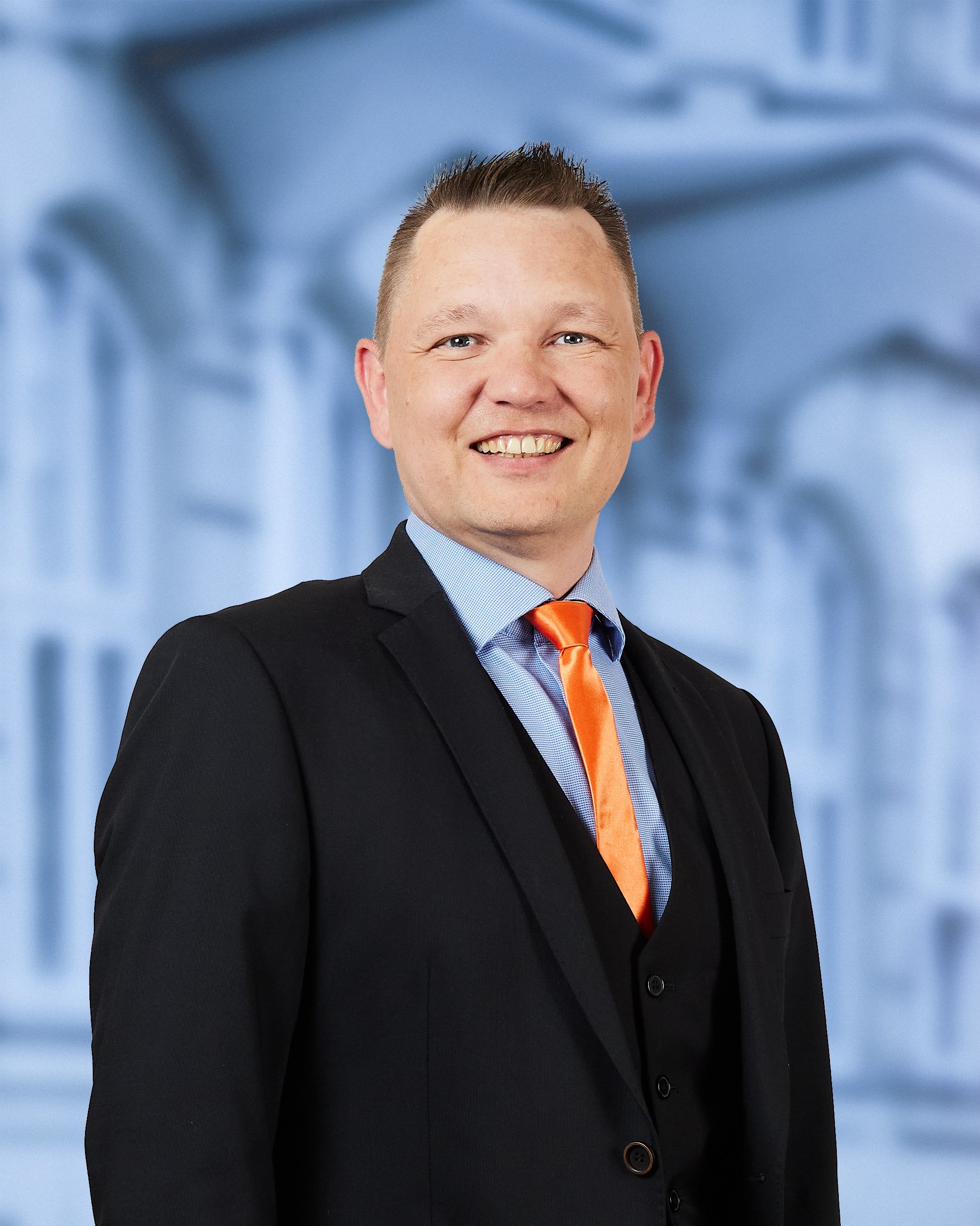 Et portræt af en mand iført sort jakkesæt, blå skjorte og orange slips, smiling mod kameraet med en blå og grå baggrund.
