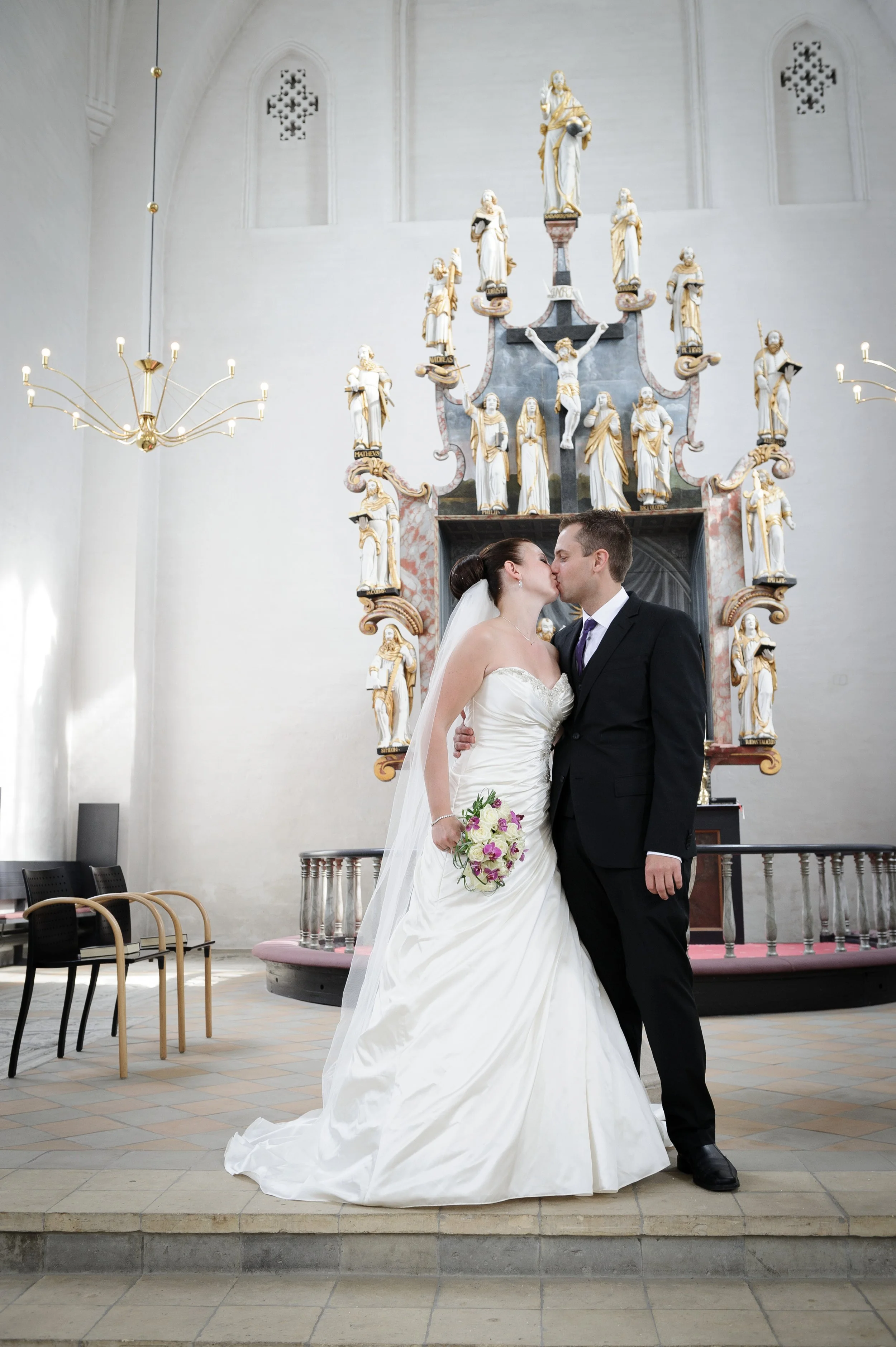 Et brudepar kysser i en kirke, hun har brudekjole og buket, han har jakkesæt, baggrunden har religiøse statuer og en alter med lysekroner.