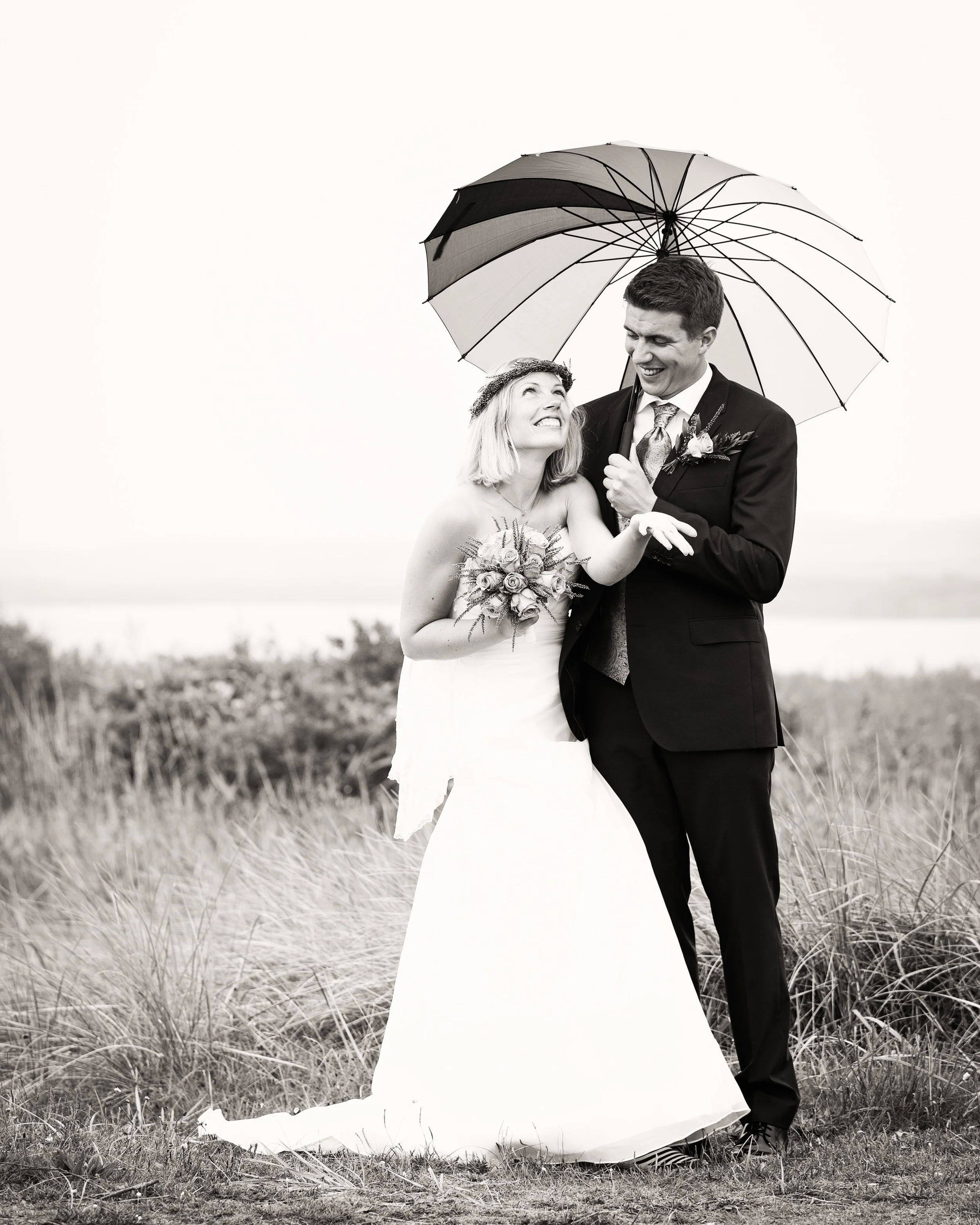 Sort-hvid foto af en glædelig brudepar, der står i en mark med græs. Bruden holder en buket blomster, og de smiler til hinanden under et sort og hvidt parasol.