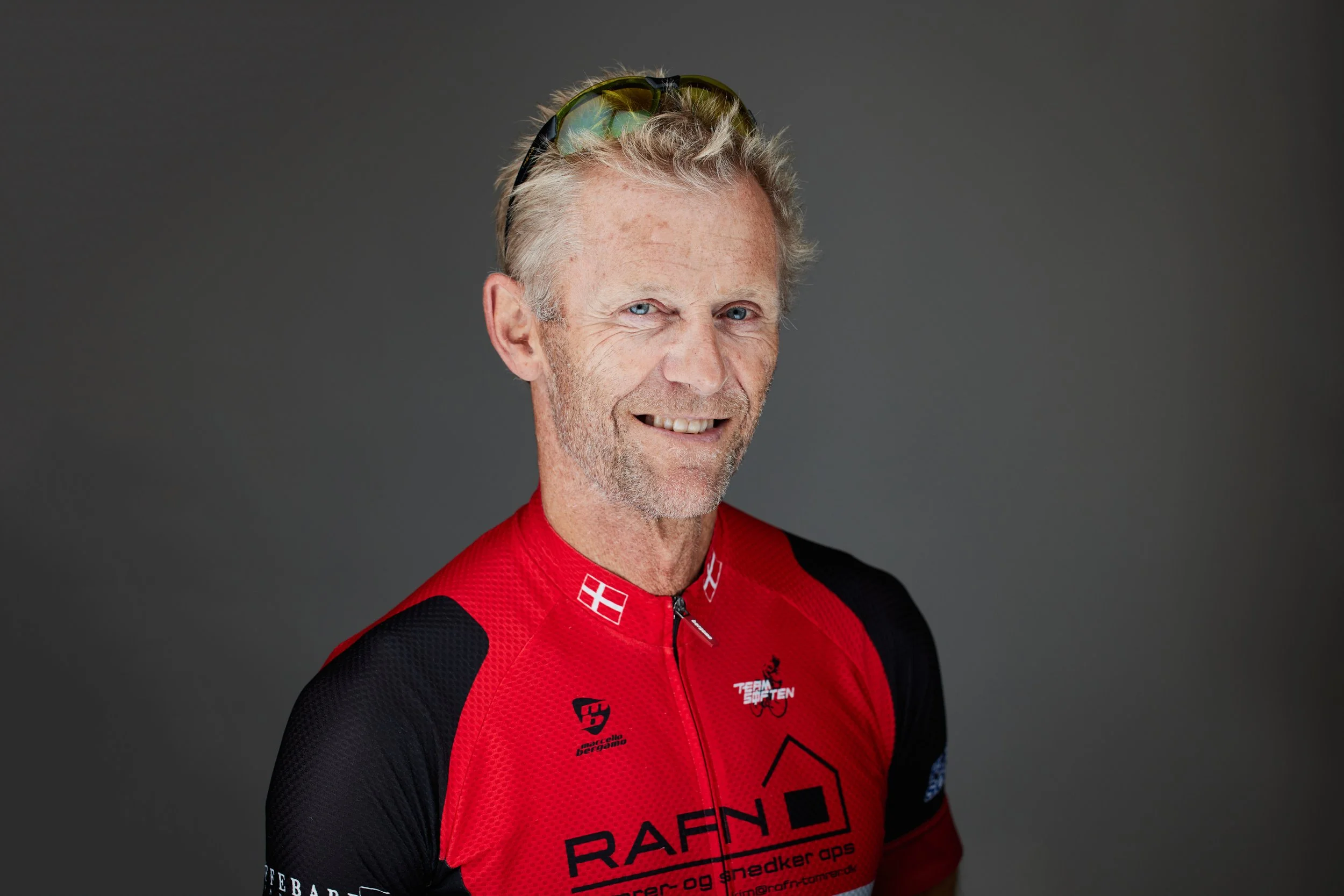 E ældre mand i en rød og sort cykeltrøje smiler mod kameraet mod en grå baggrund.