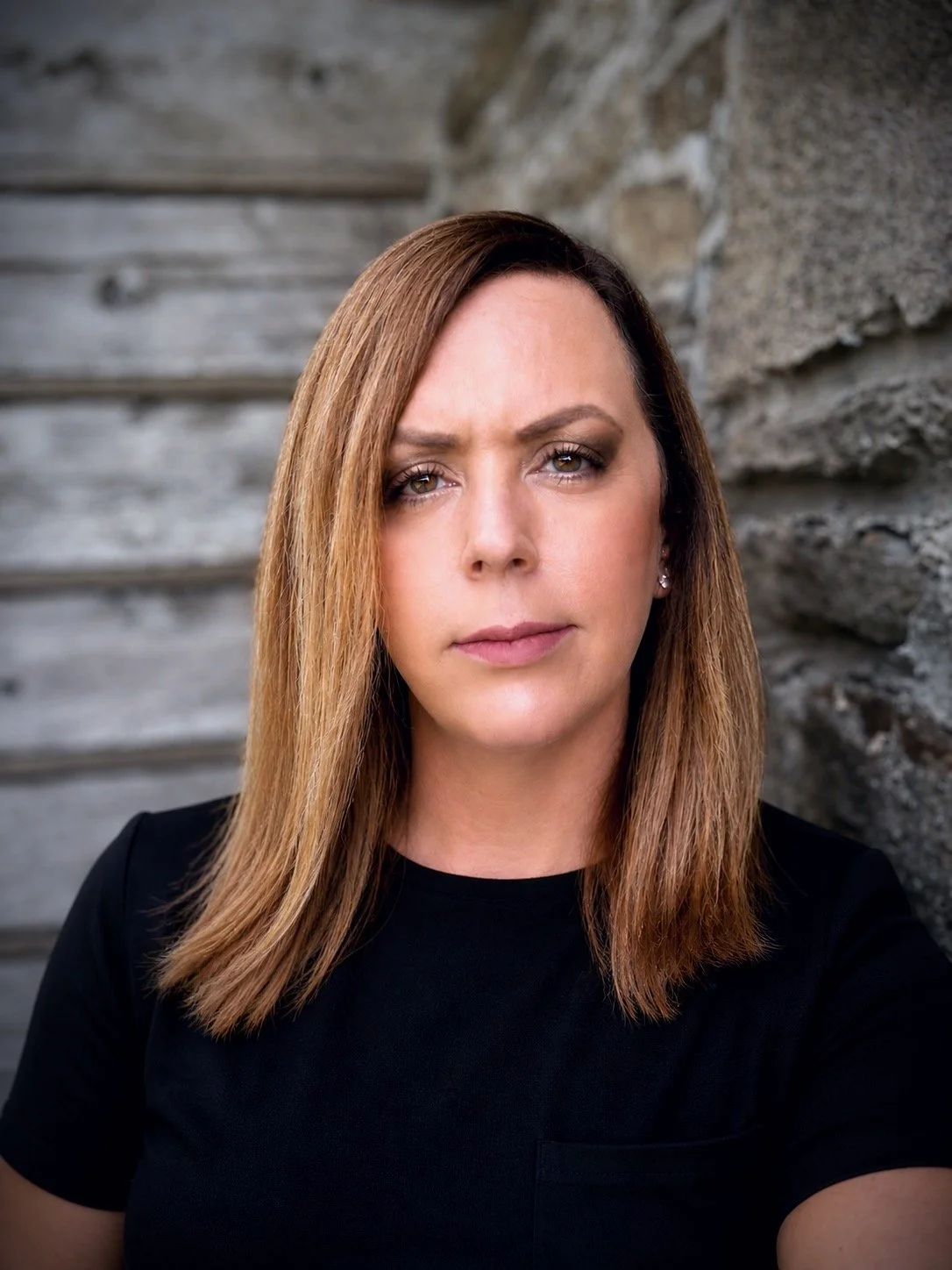 A woman named Autumn with straight, shoulder-length light brown hair, wearing a black shirt, standing outdoors against a background of wooden and stone structures.