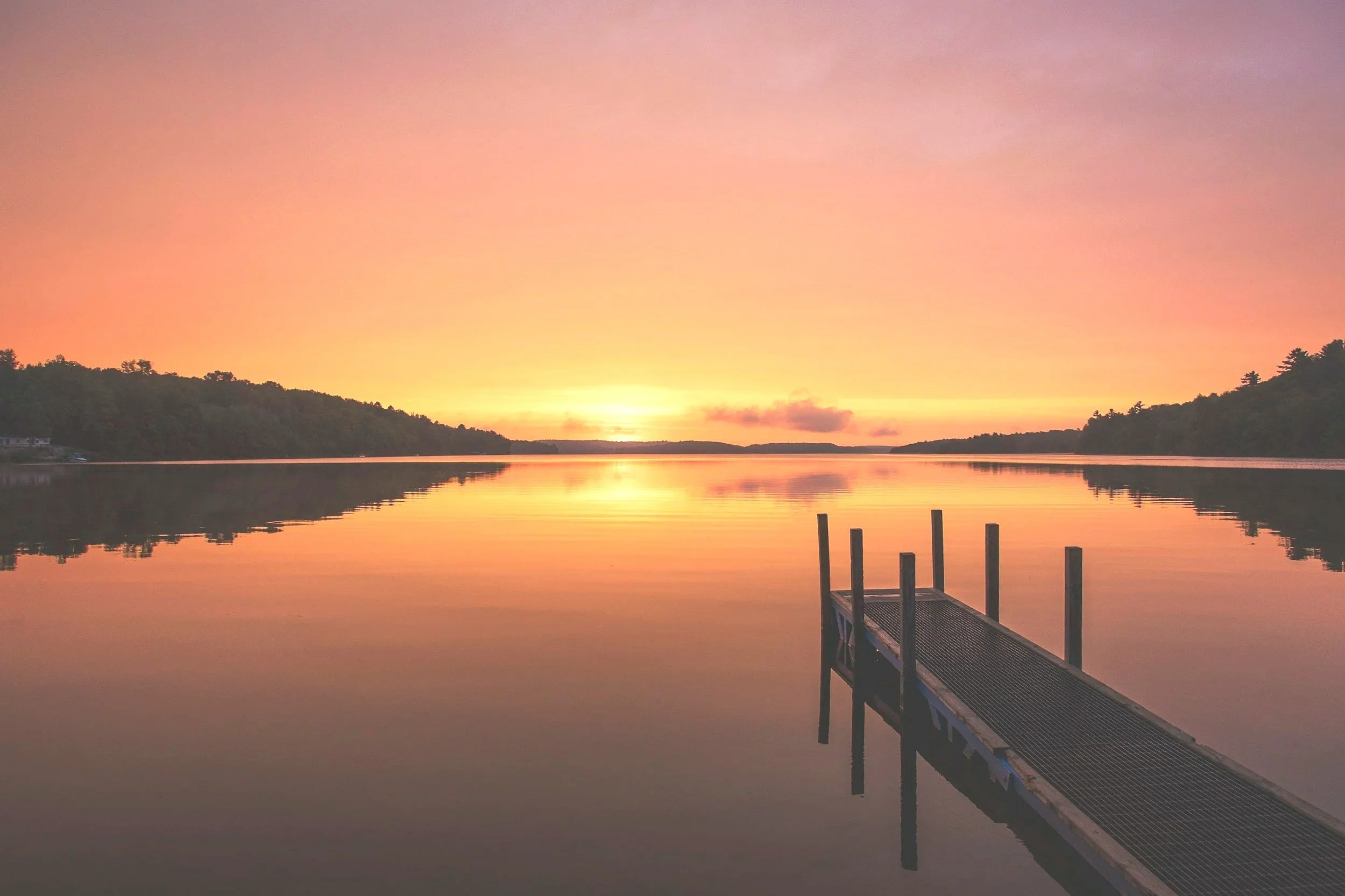 A calm lake with a dock extending into the water during sunset, with the sky painted in shades of pink, purple, and orange, and silhouettes of trees on the distant shoreline.