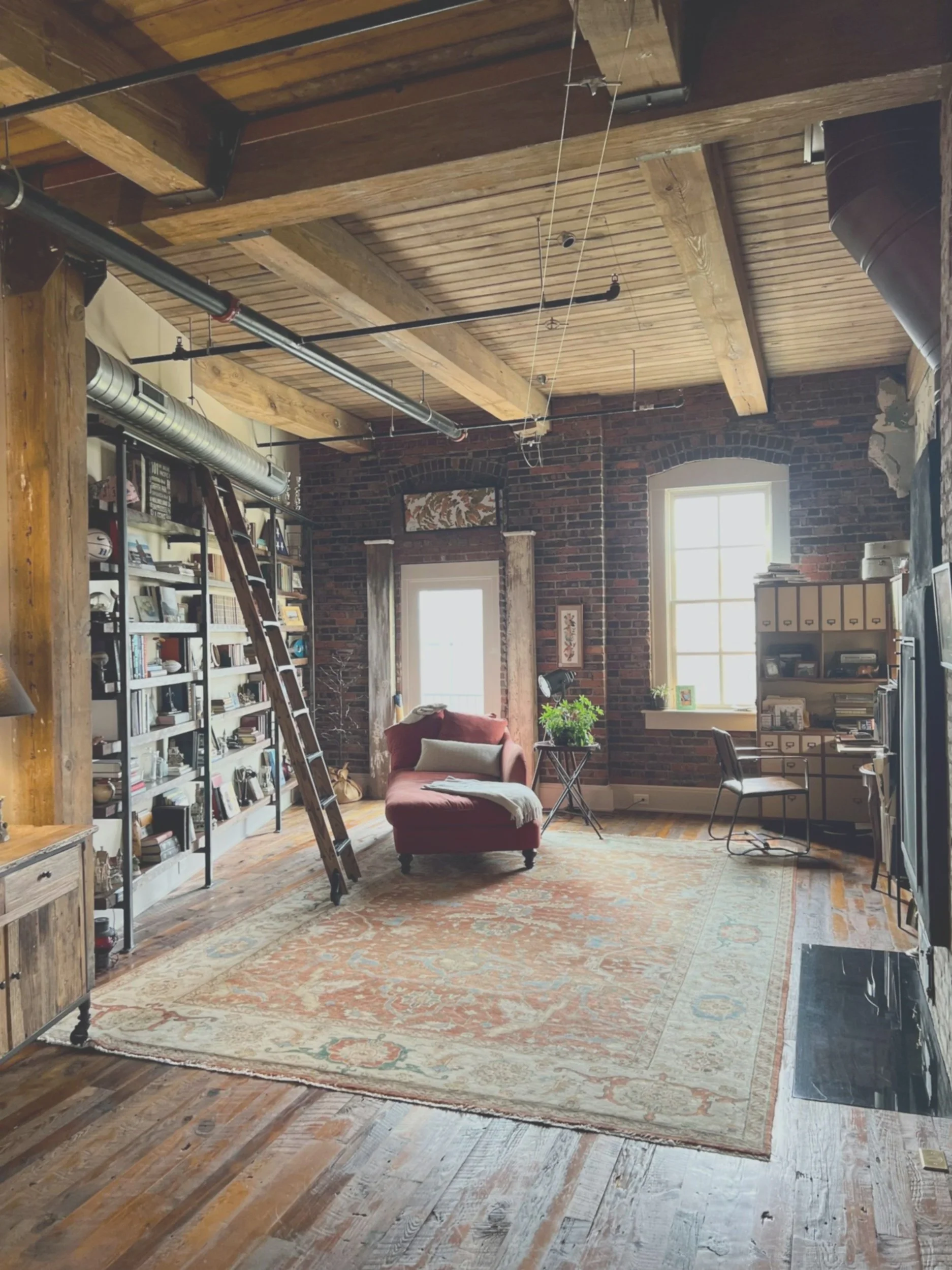 Cozy living room with brick walls, wooden beams ceiling, large windows, red armchair, bookshelf, ladder, desk, and plants.