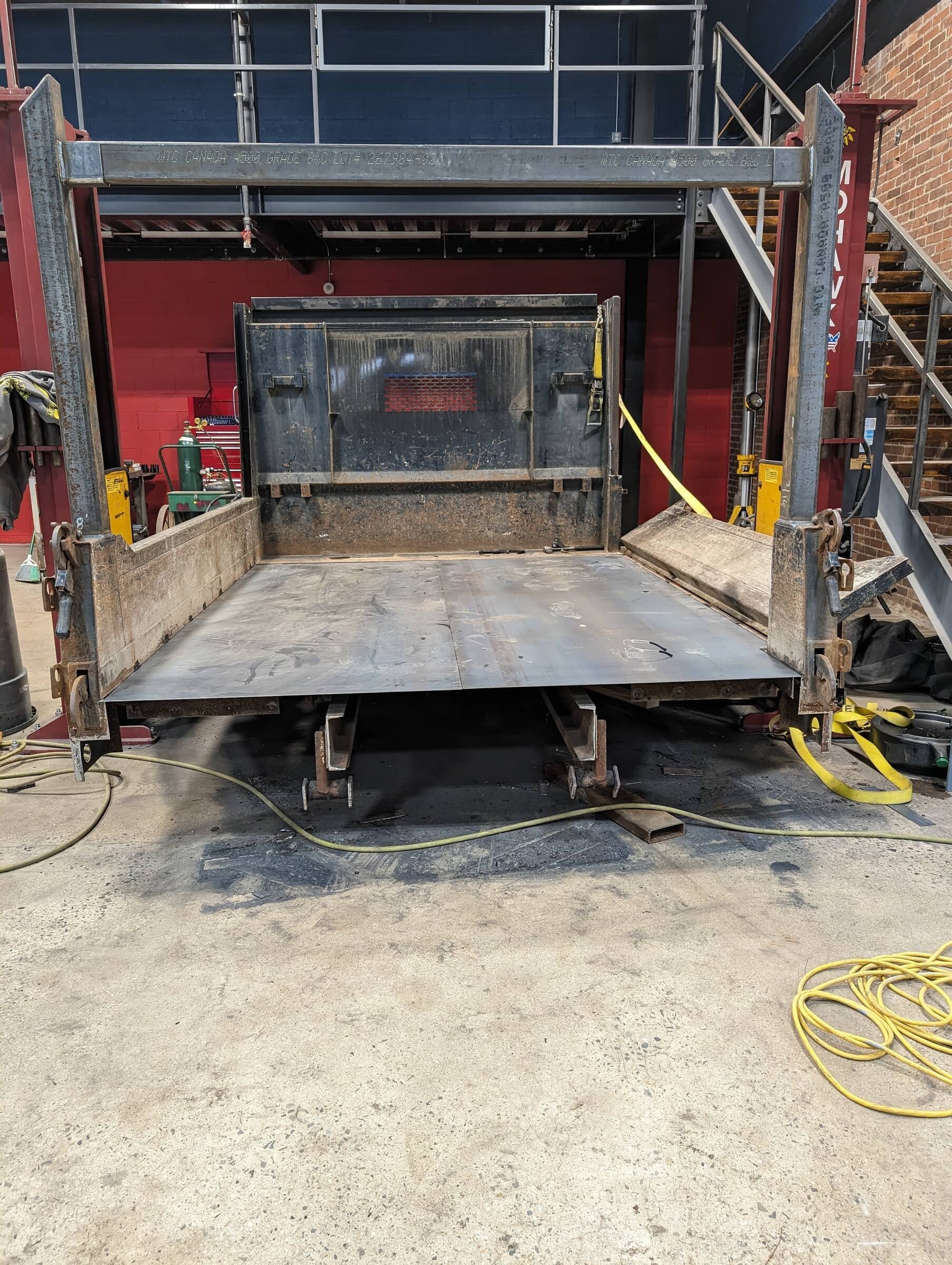 Empty industrial flatbed truck with metal sides and a black cab, parked inside a workshop with red and blue walls, welding equipment, and yellow extension cords.