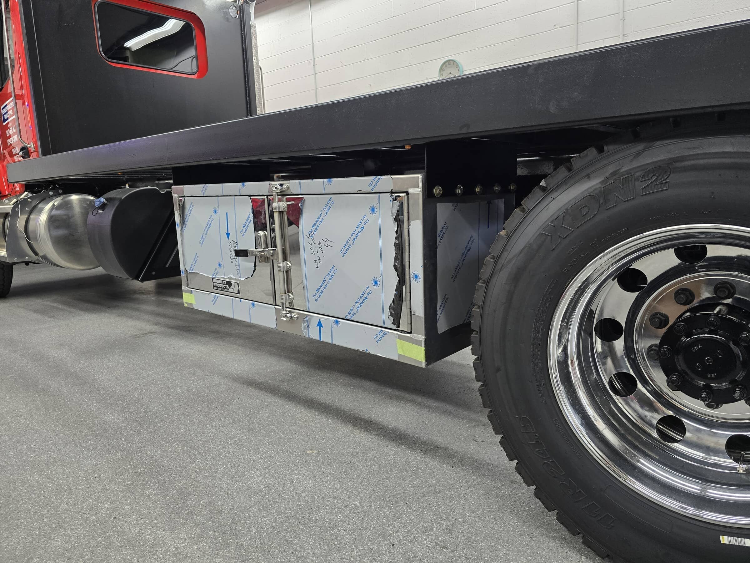 Close-up of a vehicle's side showing a black platform, metallic equipment, and a large black tire with a chrome rim inside a garage.