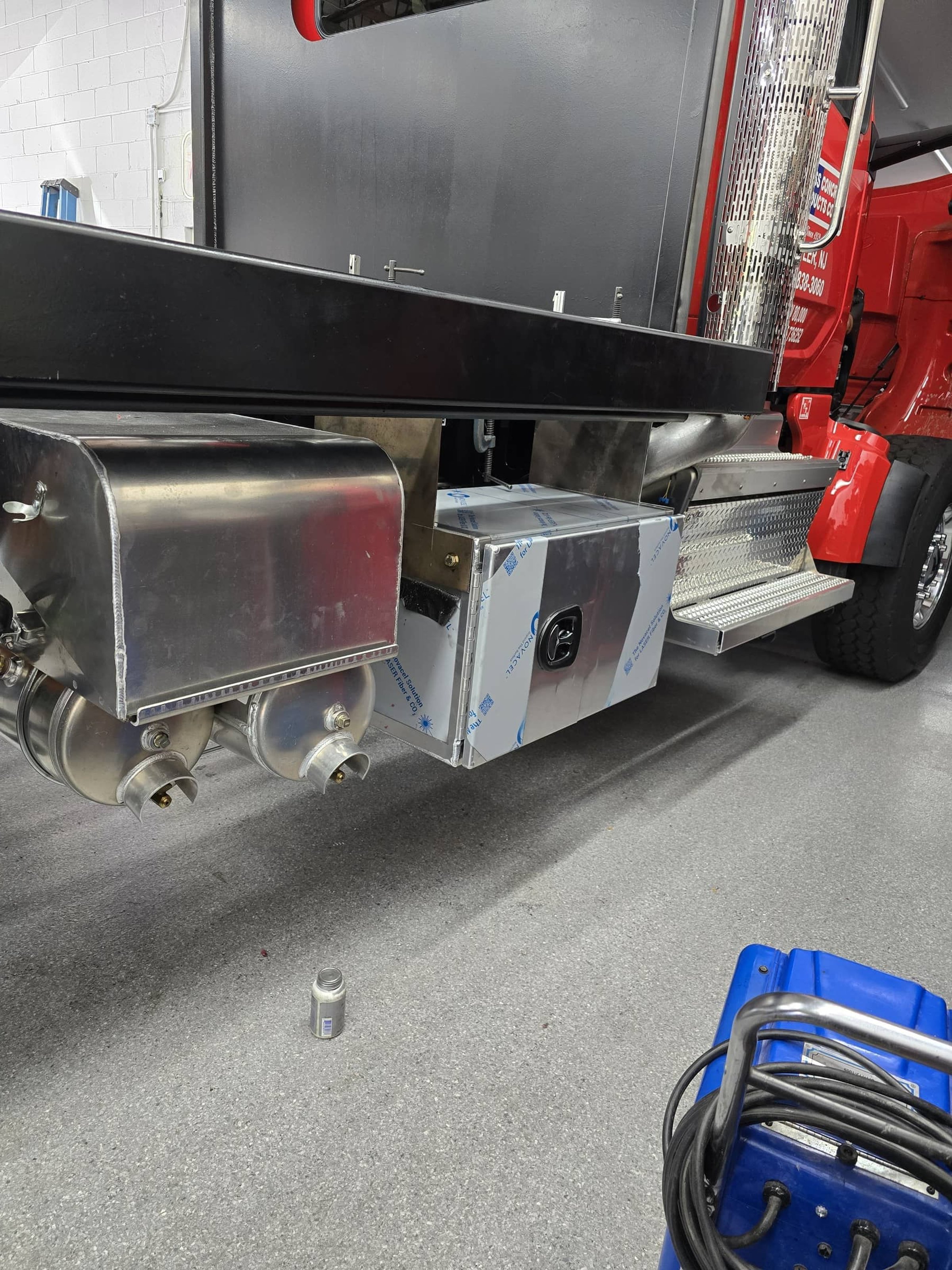 Close-up of the underside of a red truck with metallic equipment, pipes, and components installed on its side. There is a small blue tool box with cables on the ground.