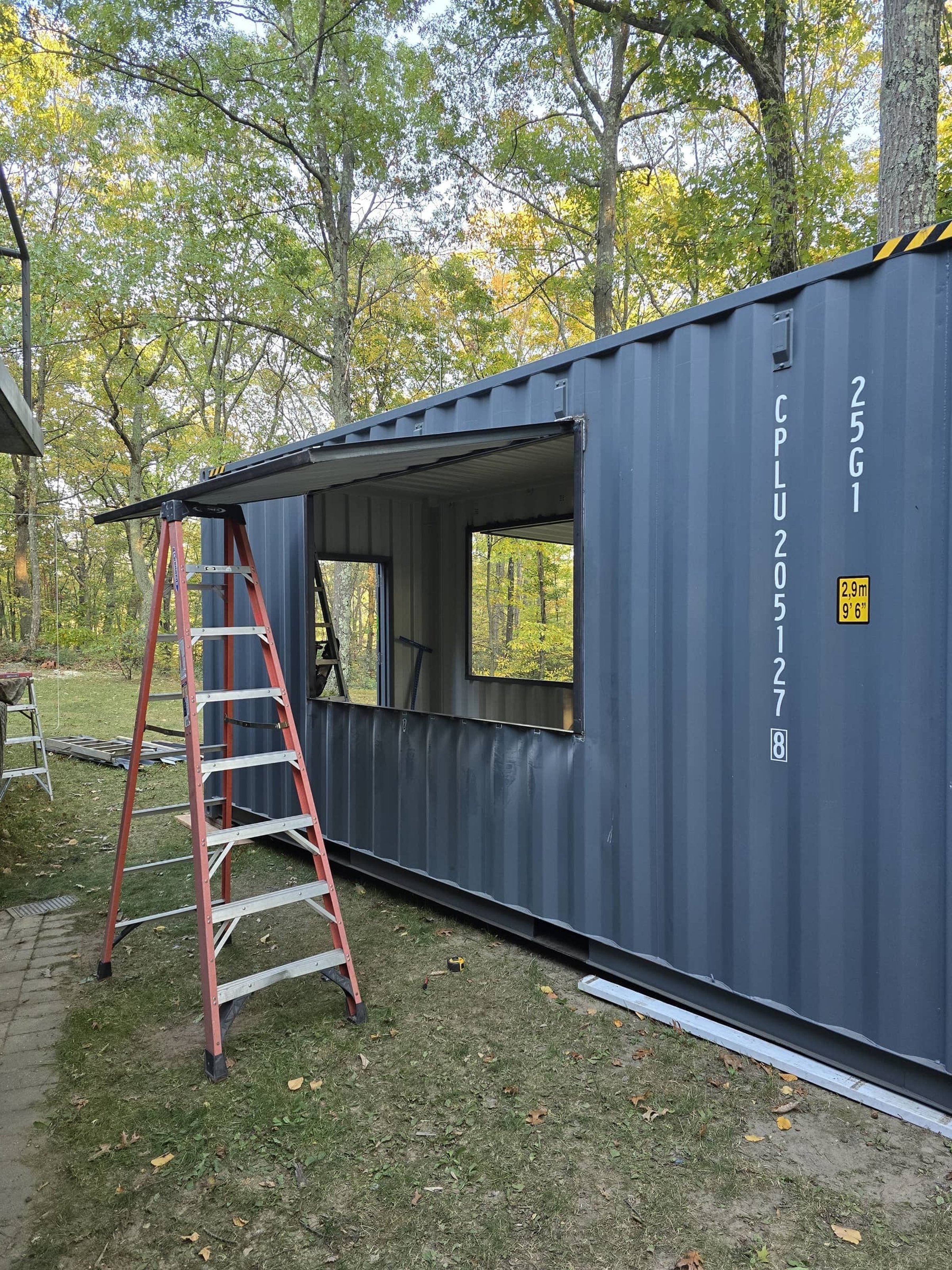 A shipping container with a large window cut out is being converted into a structure, with ladders and tools nearby, set outdoors among trees.