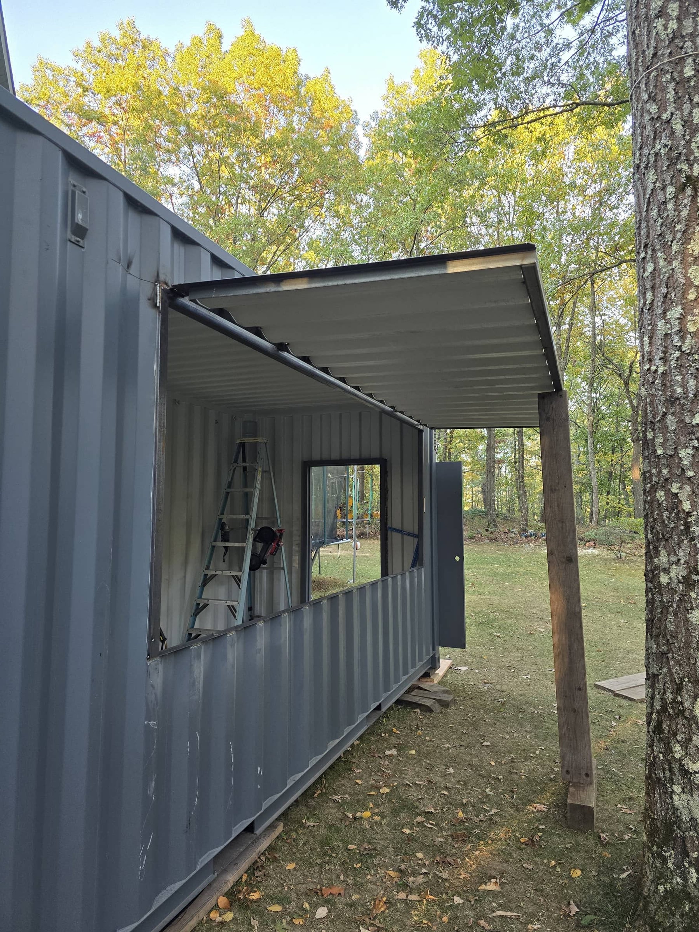 Steel shipping container with open side, window frame, metal ladder inside, and a makeshift awning supported by wooden posts in a wooded area with trees and fallen leaves.