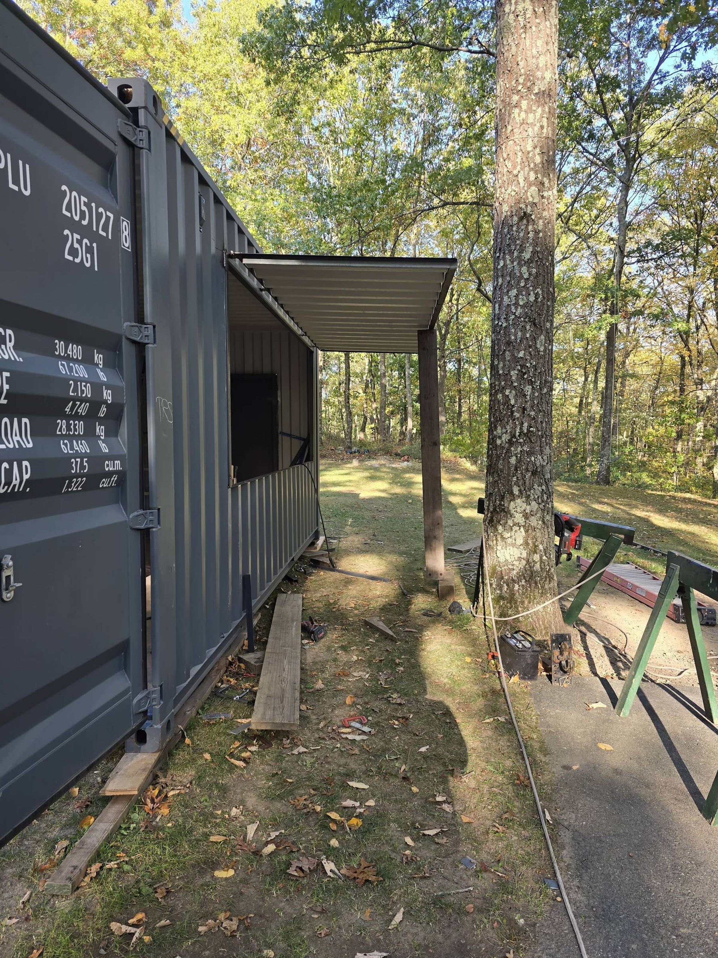 Side view of a gray portable container with open side door, set in a wooded outdoor area, with construction tools and equipment nearby.