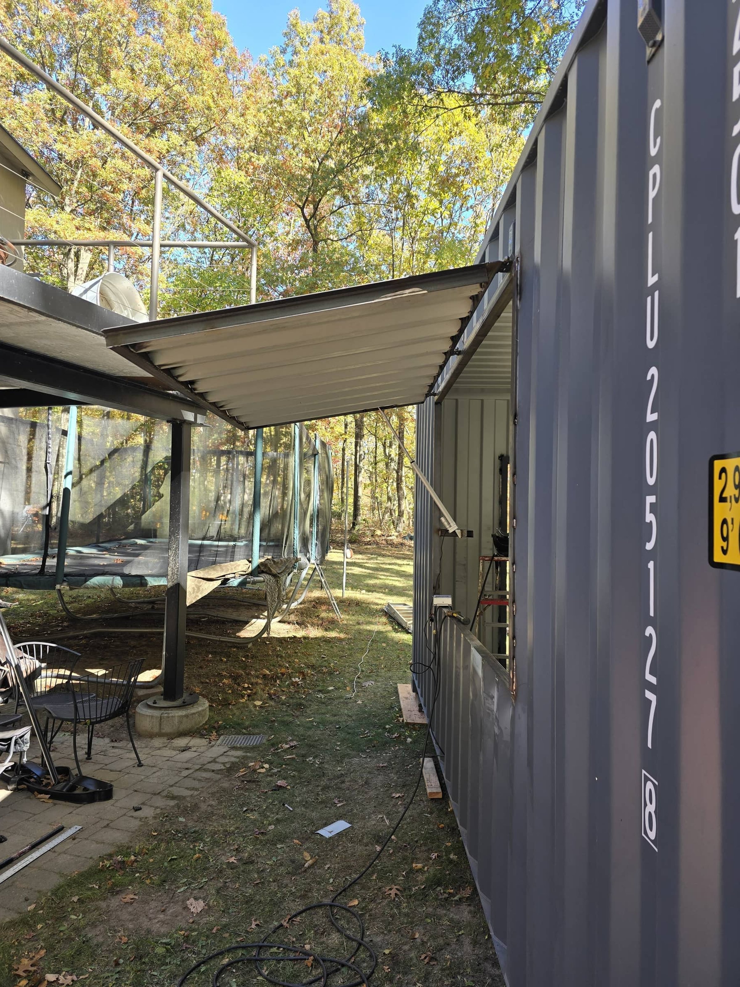 A gray shipping container with a window opening and a metal awning extension, set on a grassy outdoor area with trees, a trampoline, and patio chairs in the background.