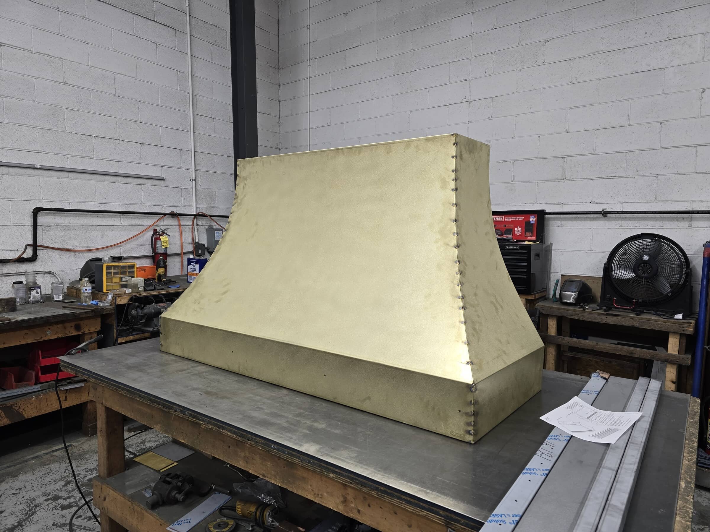 A large metal structure, possibly a part of an industrial or aerospace project, sits on a workbench in a workshop. The structure is gold-colored and has a complex geometric shape with beveled edges and visible rivets along the seams.