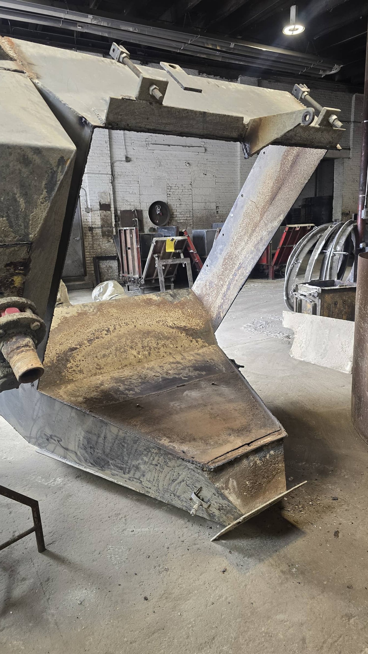 A large, rusty industrial metal scoop attached to a machinery, in a warehouse or workshop setting with brick walls and metal equipment in the background.