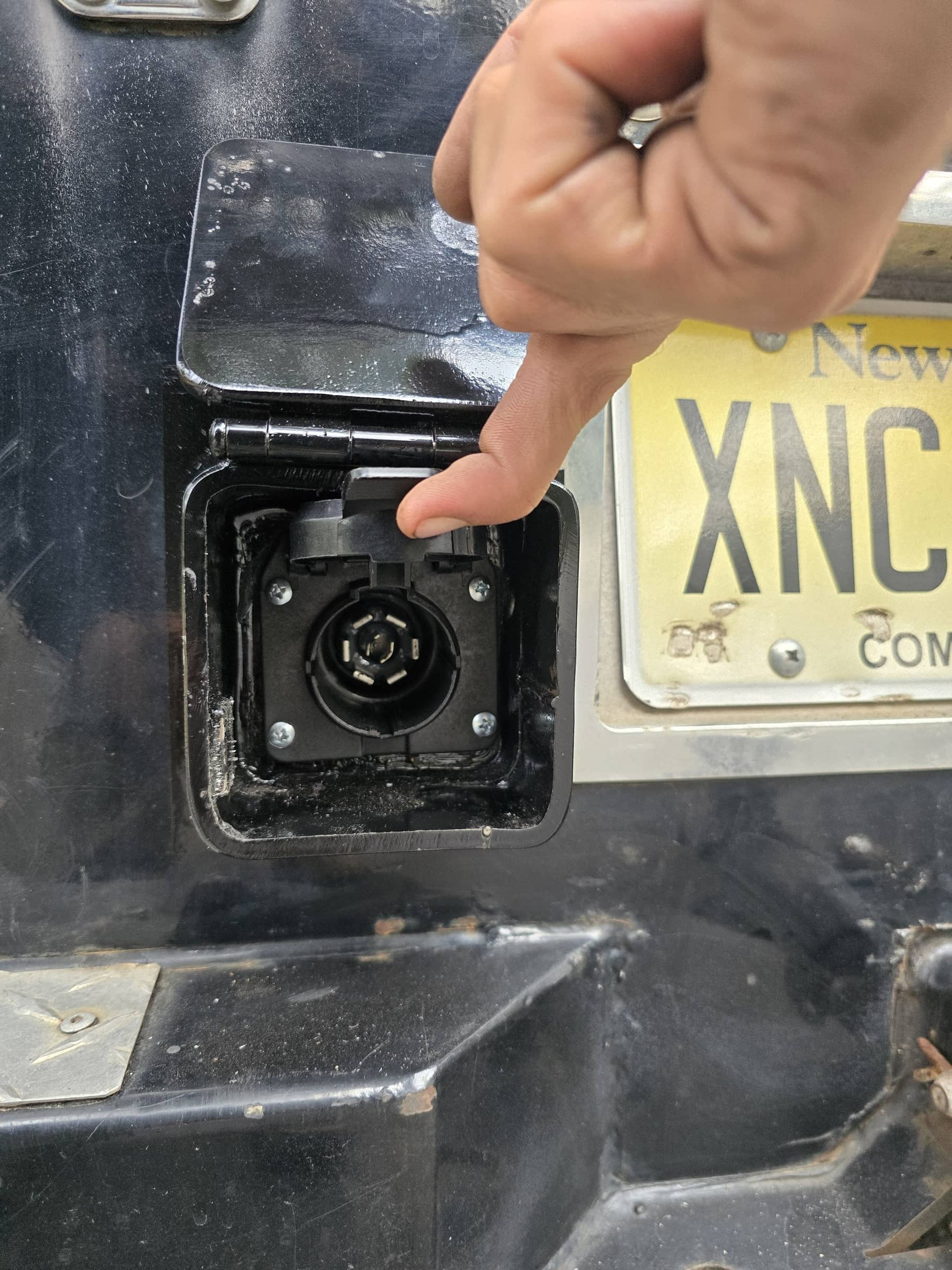 A person holding a metal cover open to reveal an electrical outlet socket, with a license plate partially visible in the background.