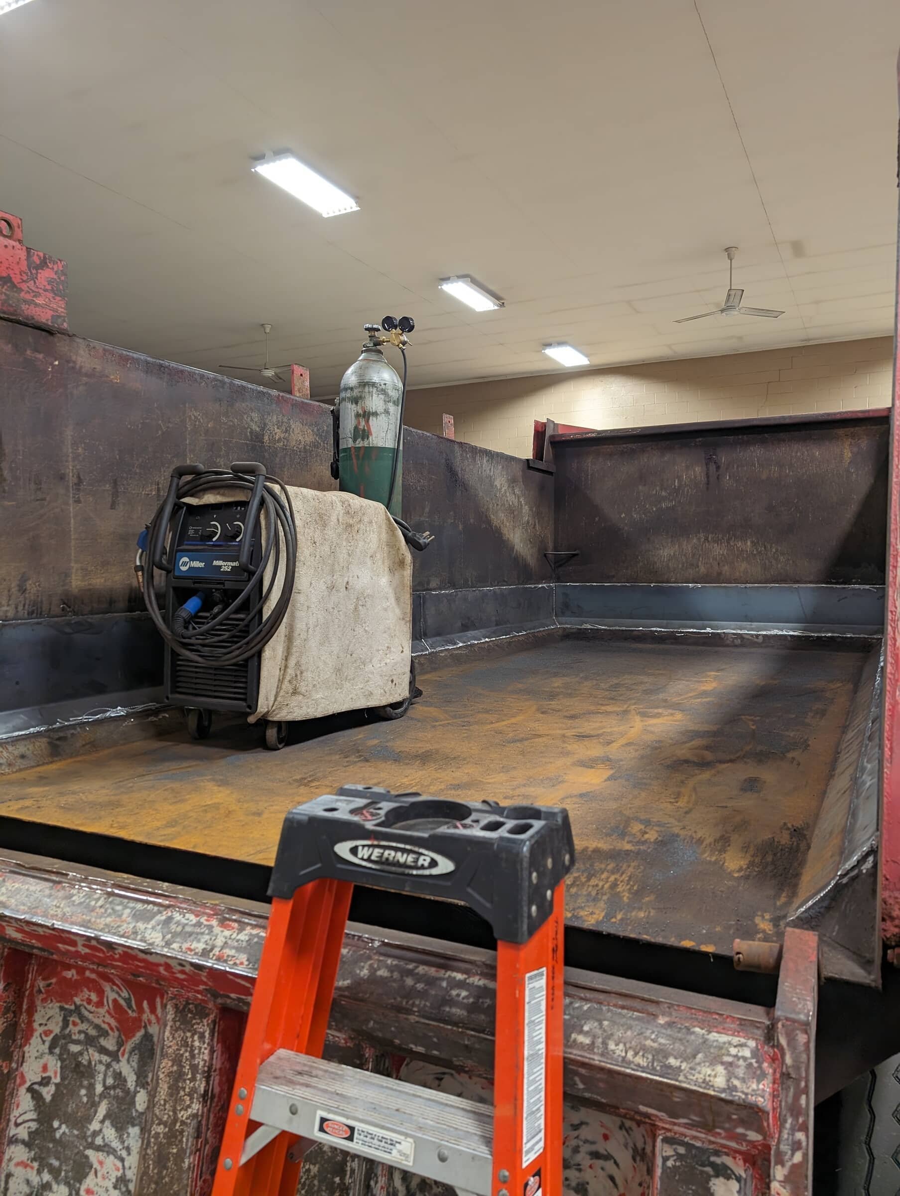 Industrial workspace with welding equipment, a scissor lift, and a work surface with rust and soot, illuminated by ceiling lights.