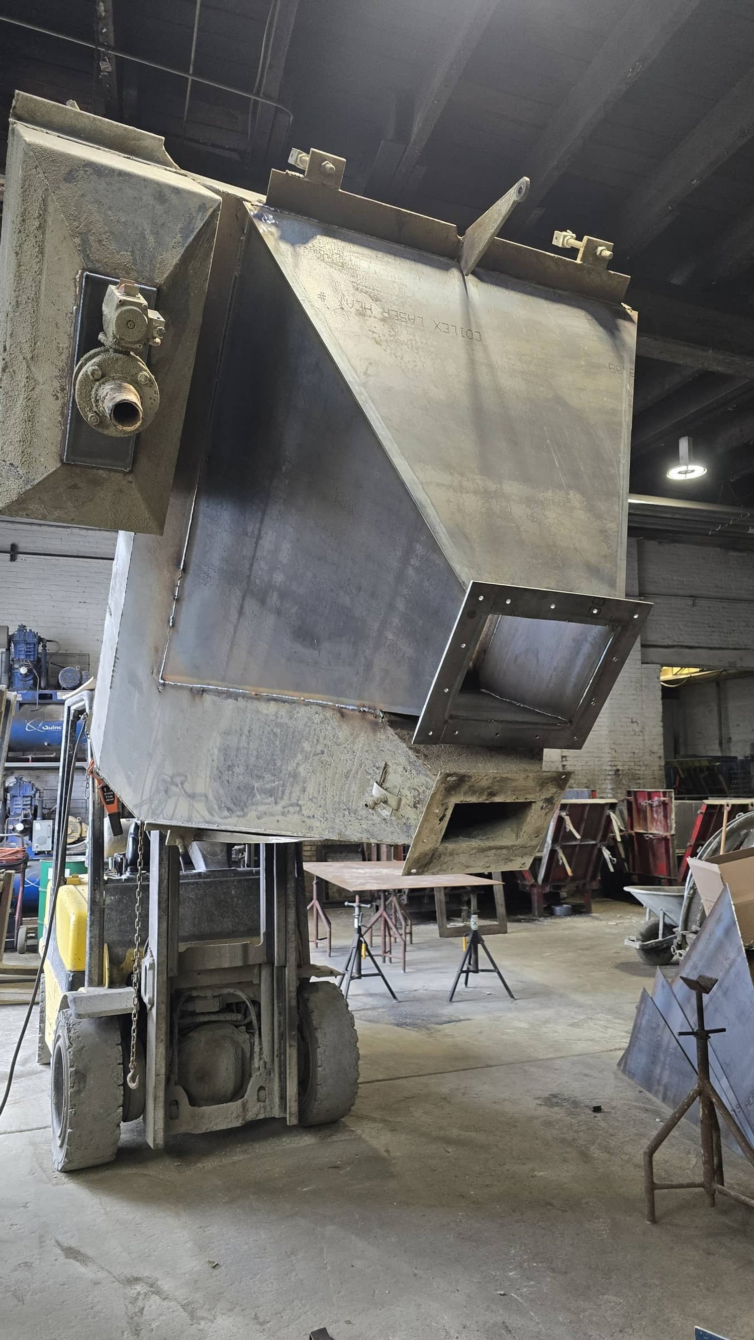 Large metal industrial tank or container being assembled or repaired in a warehouse, with various tools and equipment in the background.