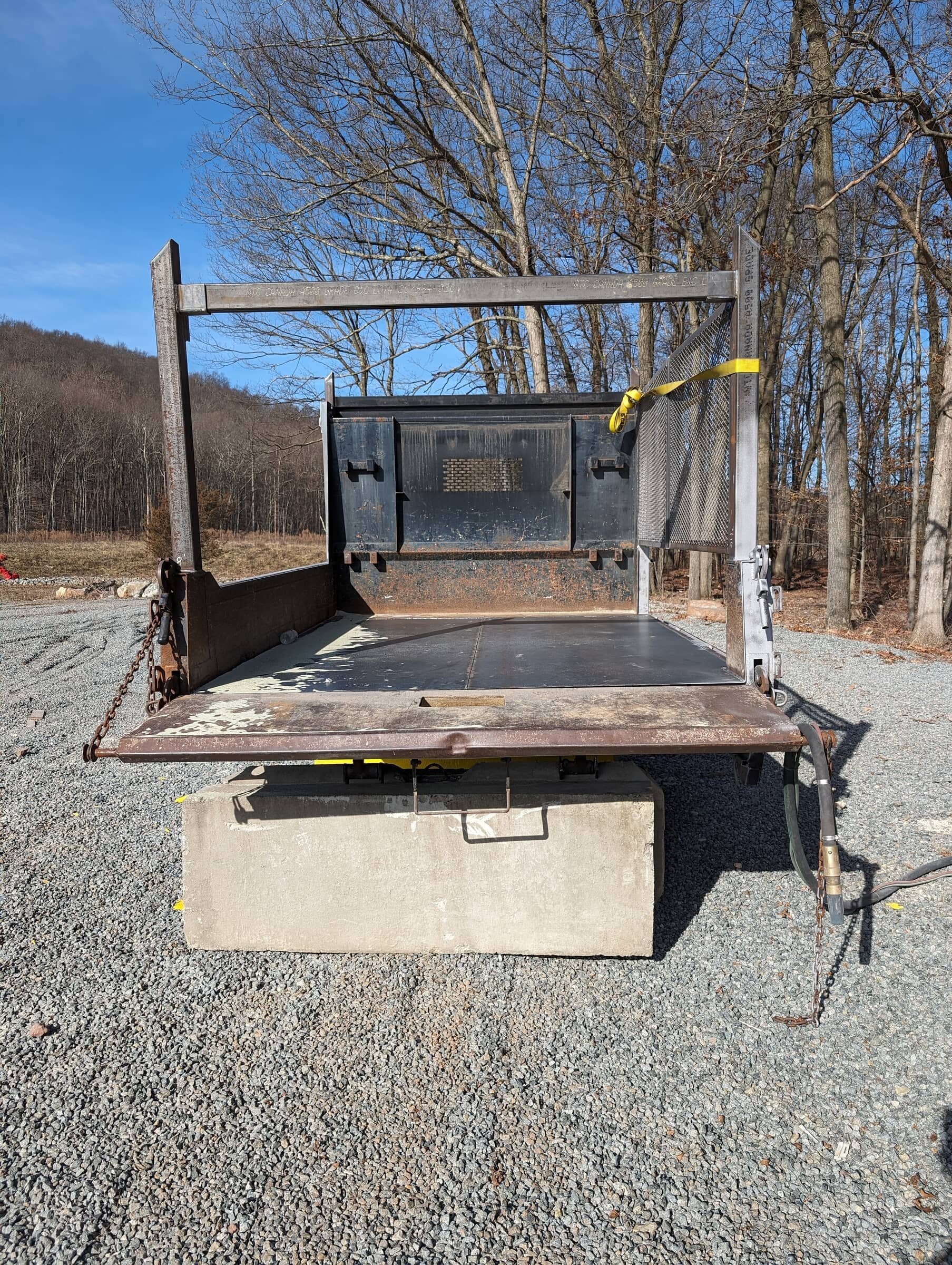 A flatbed utility trailer with a metal bed, rusted edges, chains on both sides, and a concrete block beneath. The background shows leafless trees and a gravel lot under a clear blue sky.