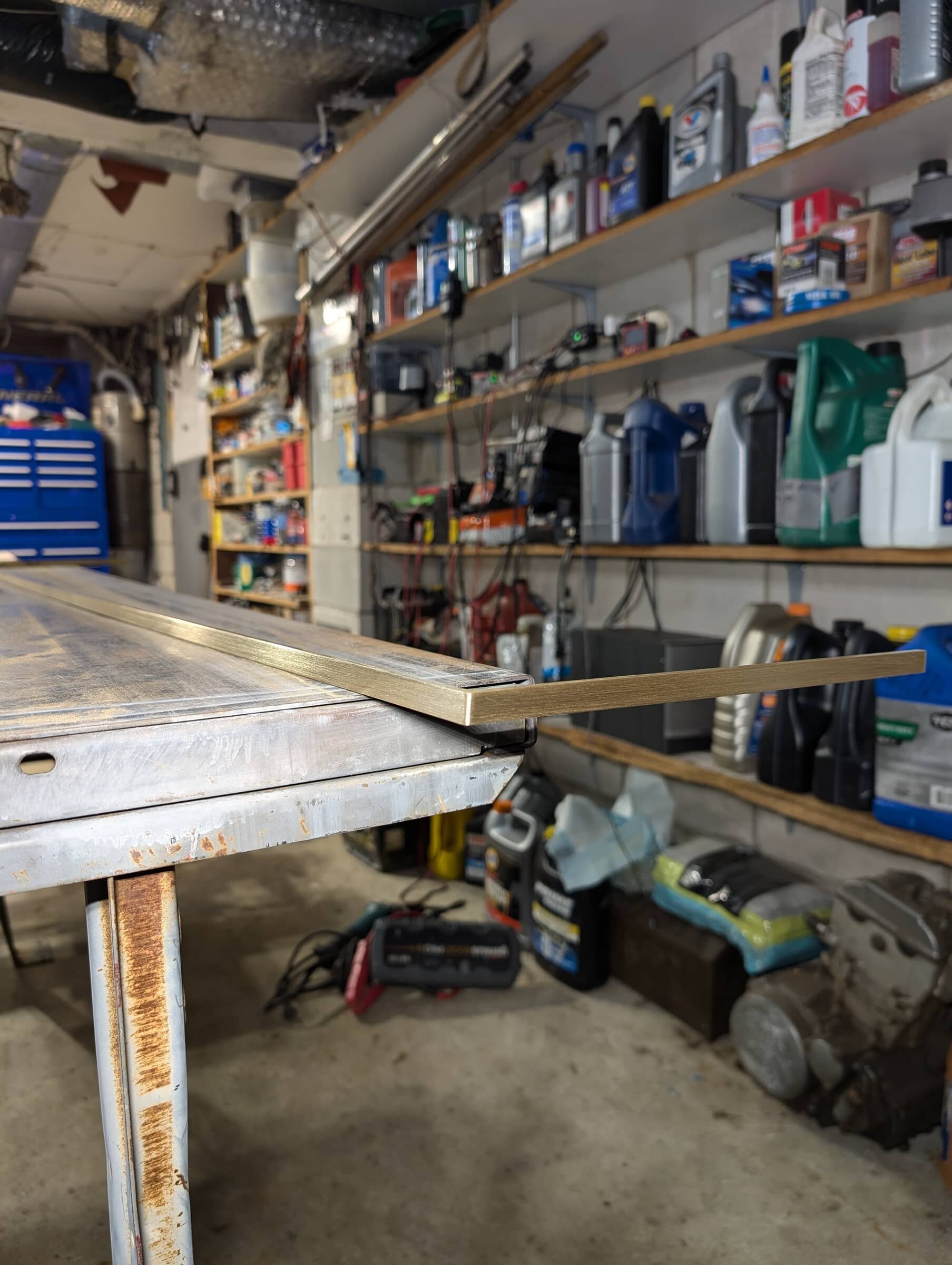 A workbench in a garage with a metal framed piece of wood extending from it, and a background of shelves stocked with bottles, cans, and tools.