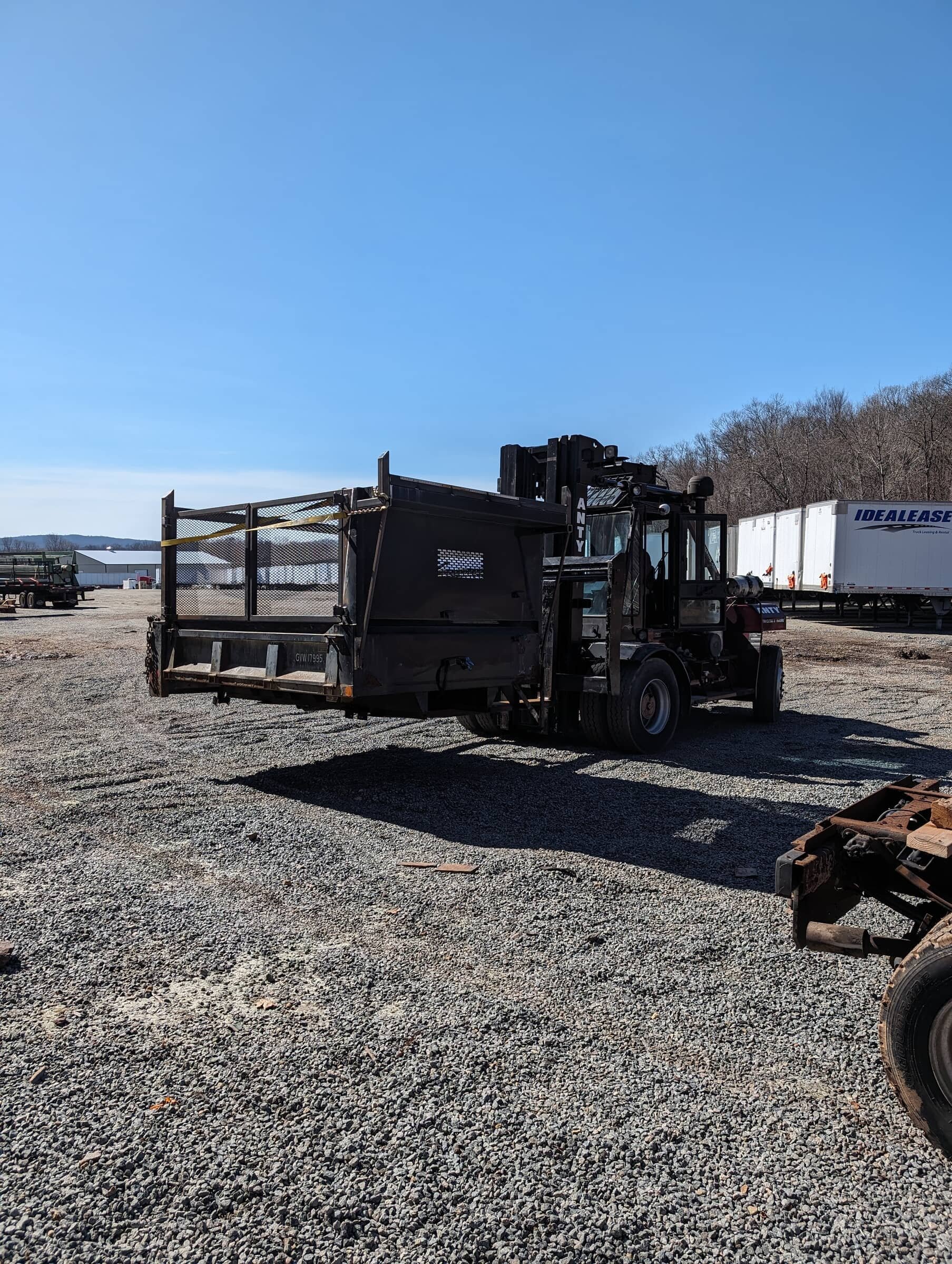 A black forklift truck on a gravel lot under a clear blue sky, with white trailers and trees in the background.
