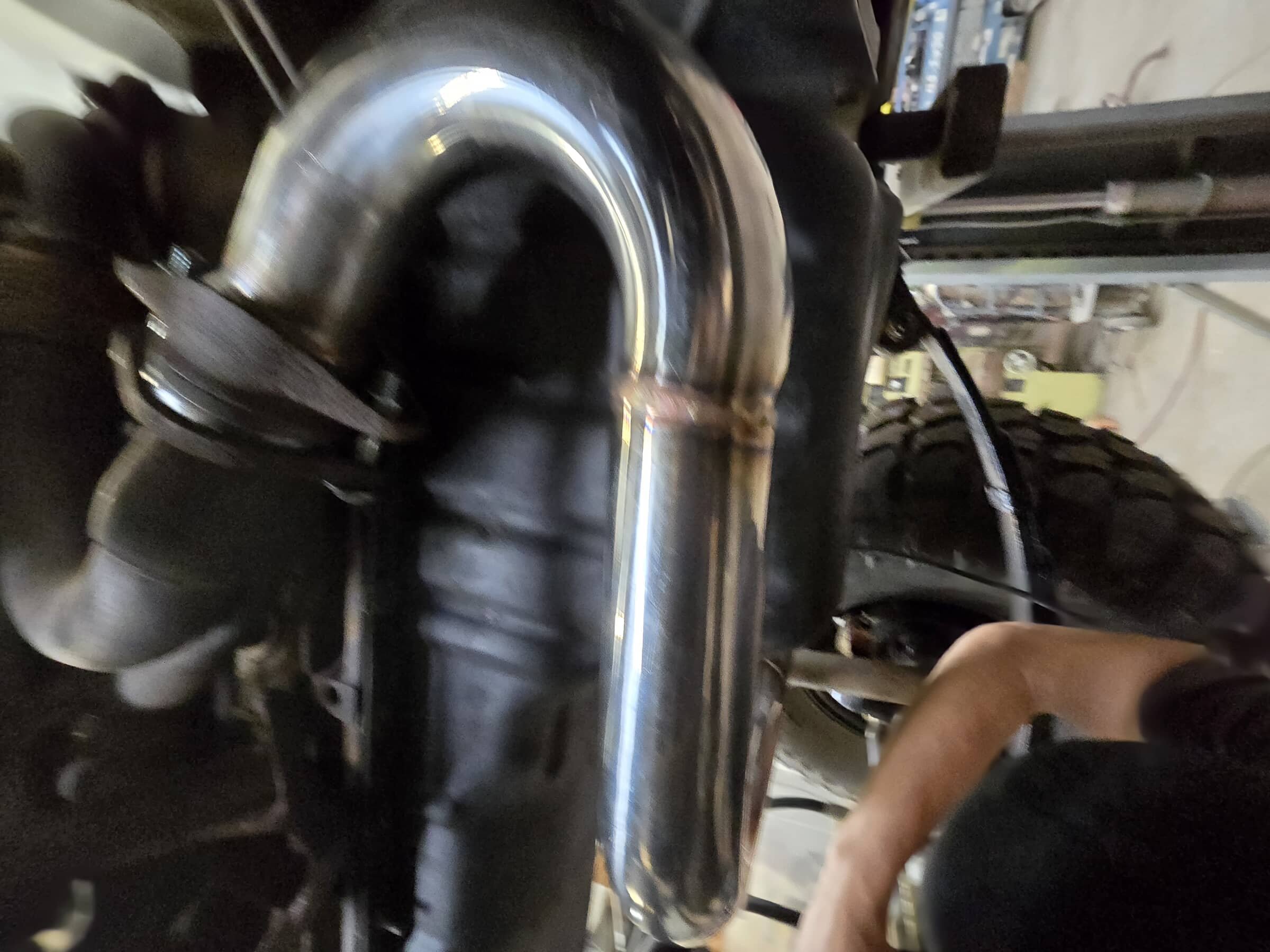Close-up of a motorcycle engine and chrome exhaust pipe, with a person’s arm visible in the bottom right corner.