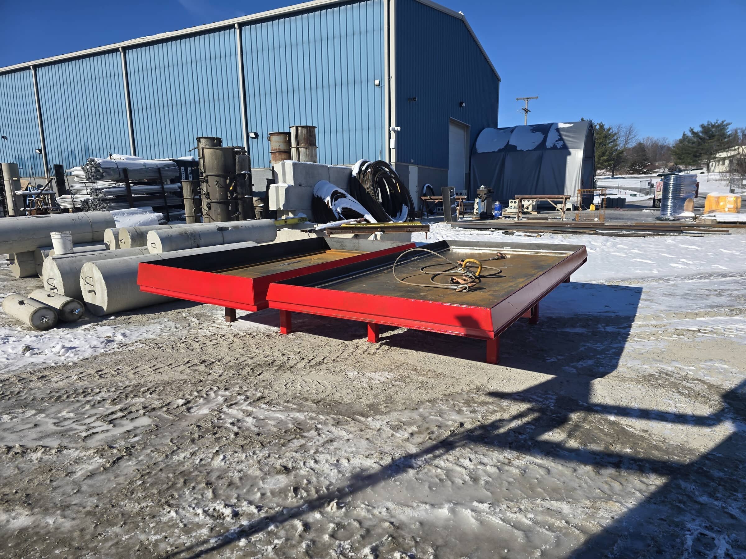 Outdoor industrial yard with large red metal platform, stacks of pipes, construction materials, and a blue building with a snow-covered ground and clear blue sky.