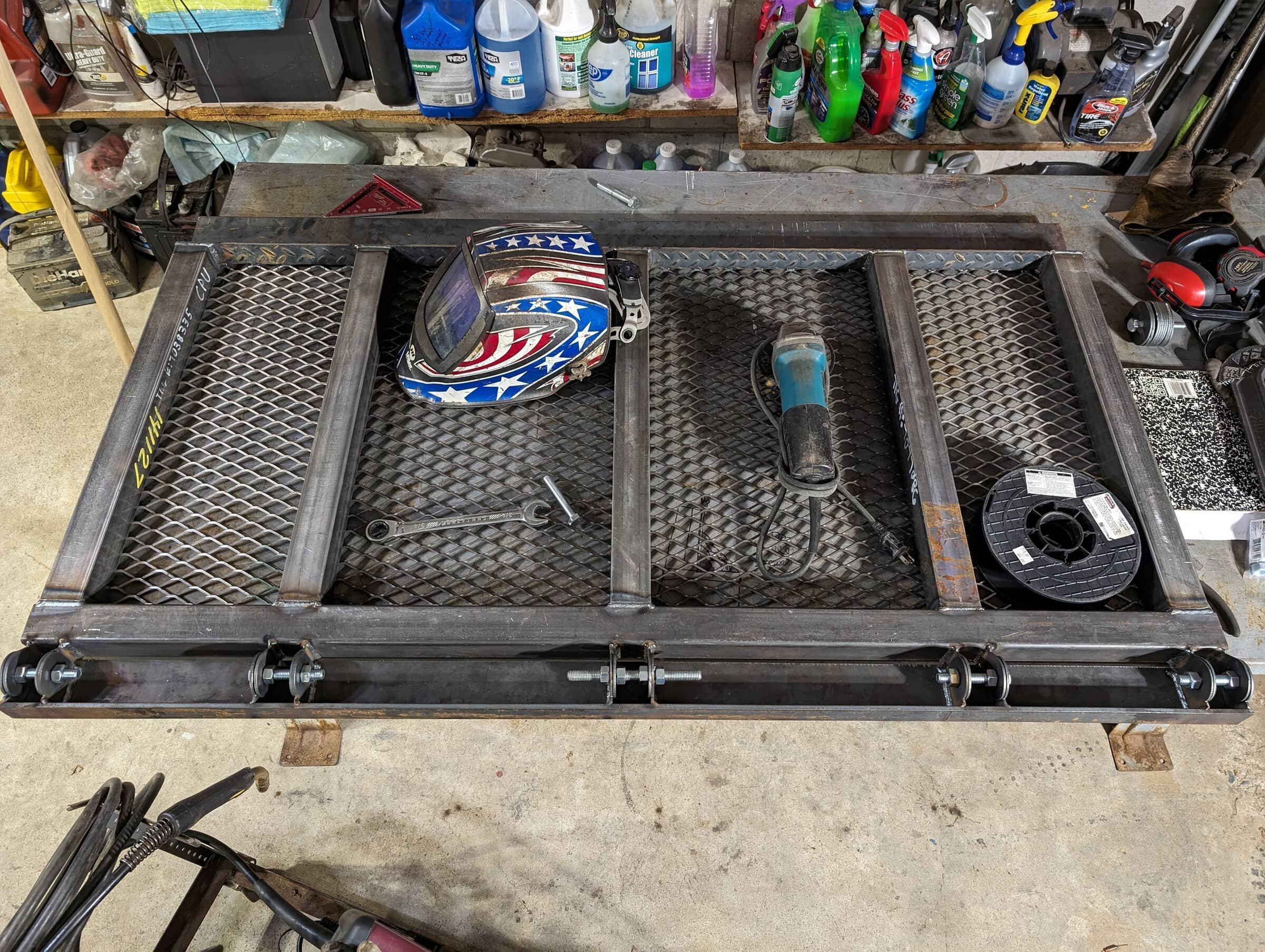 A metal work table with a welding helmet, a welding clamp, a power tool, and a spool of wire on it, in a workshop with shelves of various cleaning supplies and tools in the background.