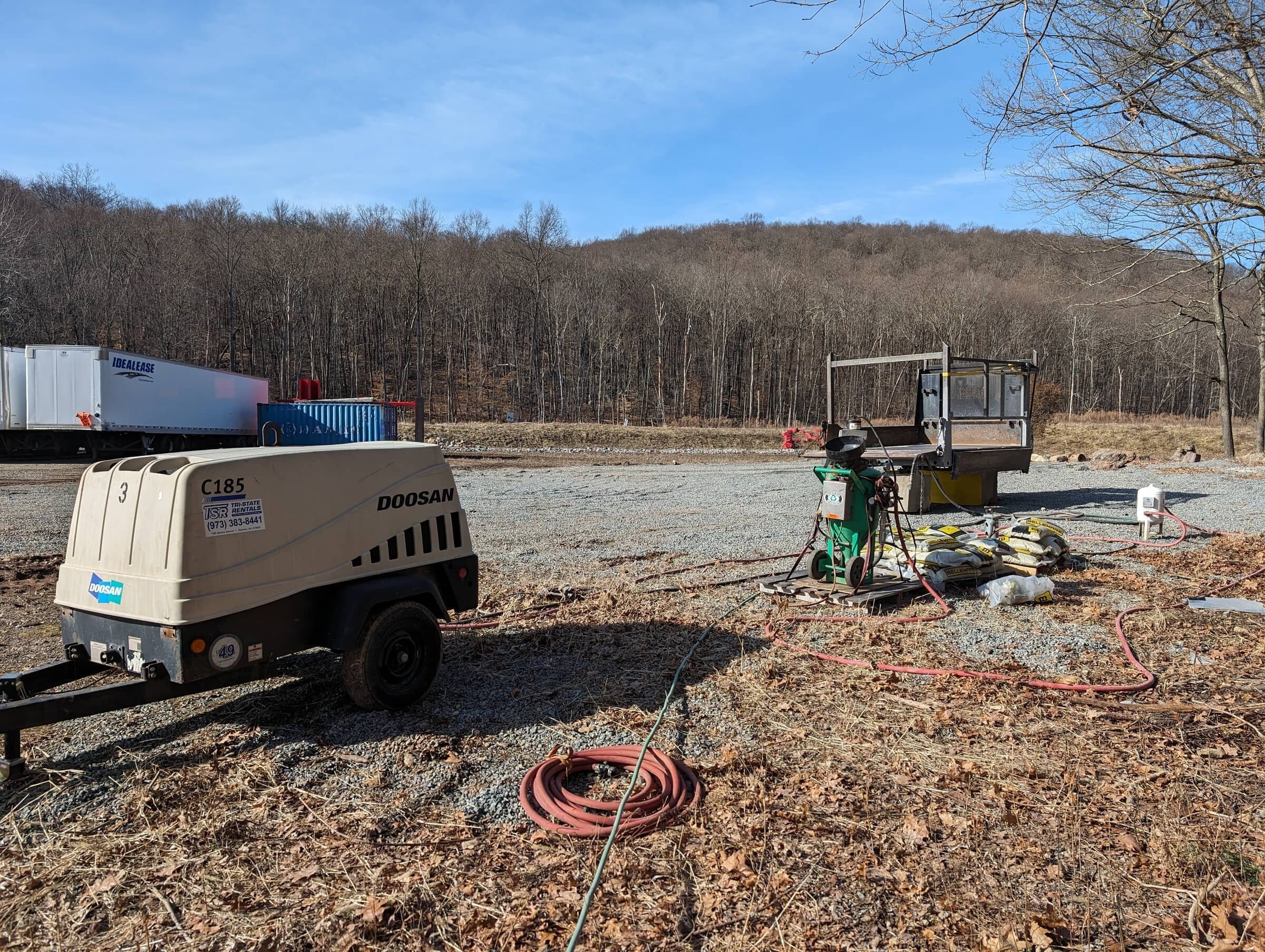 Construction site with equipment including a portable air compressor, bags of material, hoses, and a small platform, set against a background of leafless trees and a hill under a blue sky.
