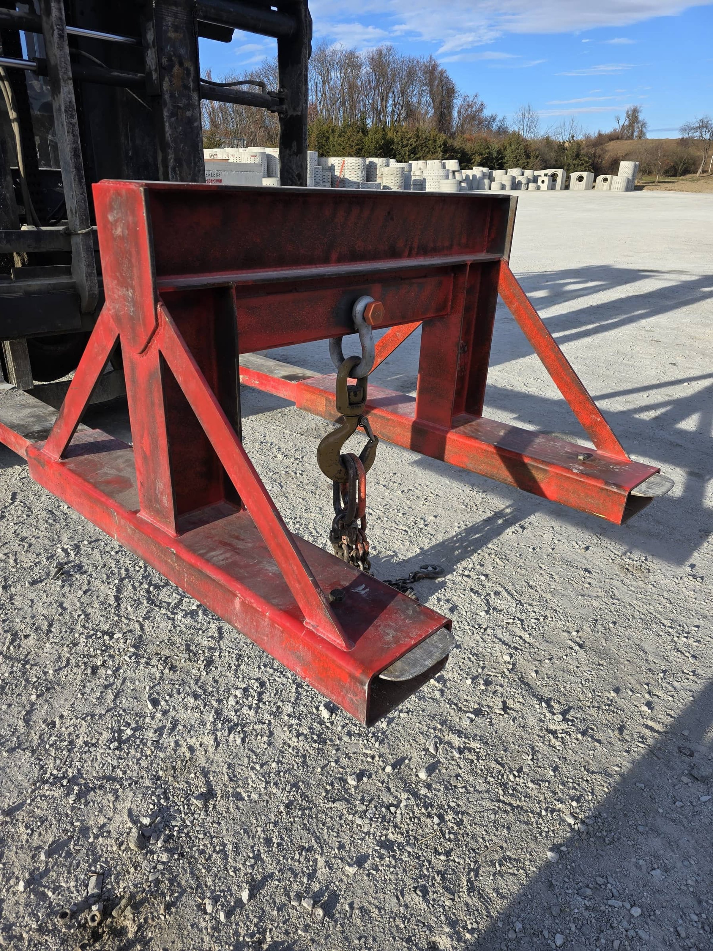 Close-up of a red steel trailer hitch with chains, set on gravel ground with pallets in the background and blue sky overhead.