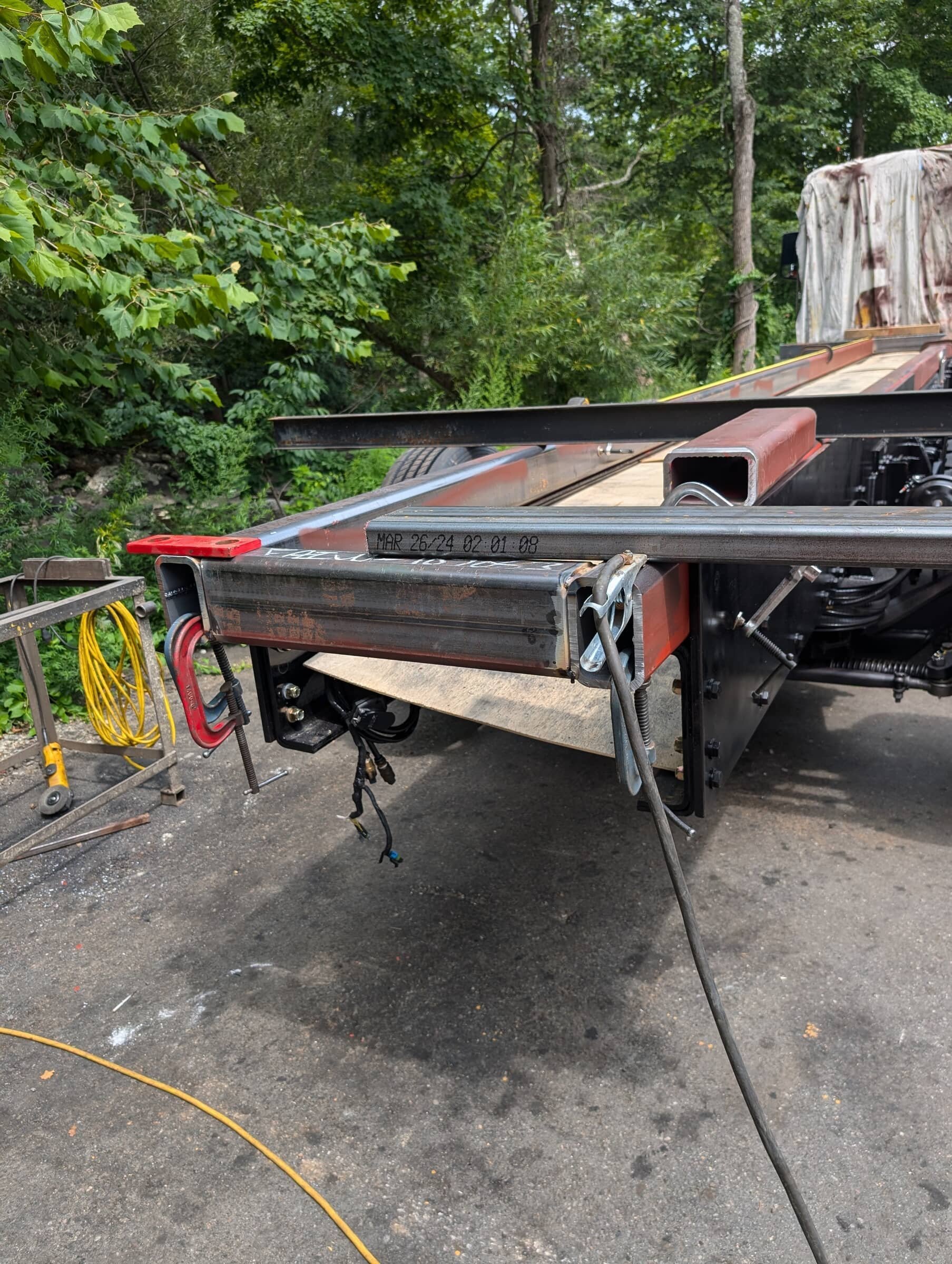 Part of a flatbed truck with metal beams and tools, green trees and a wooded area in the background.