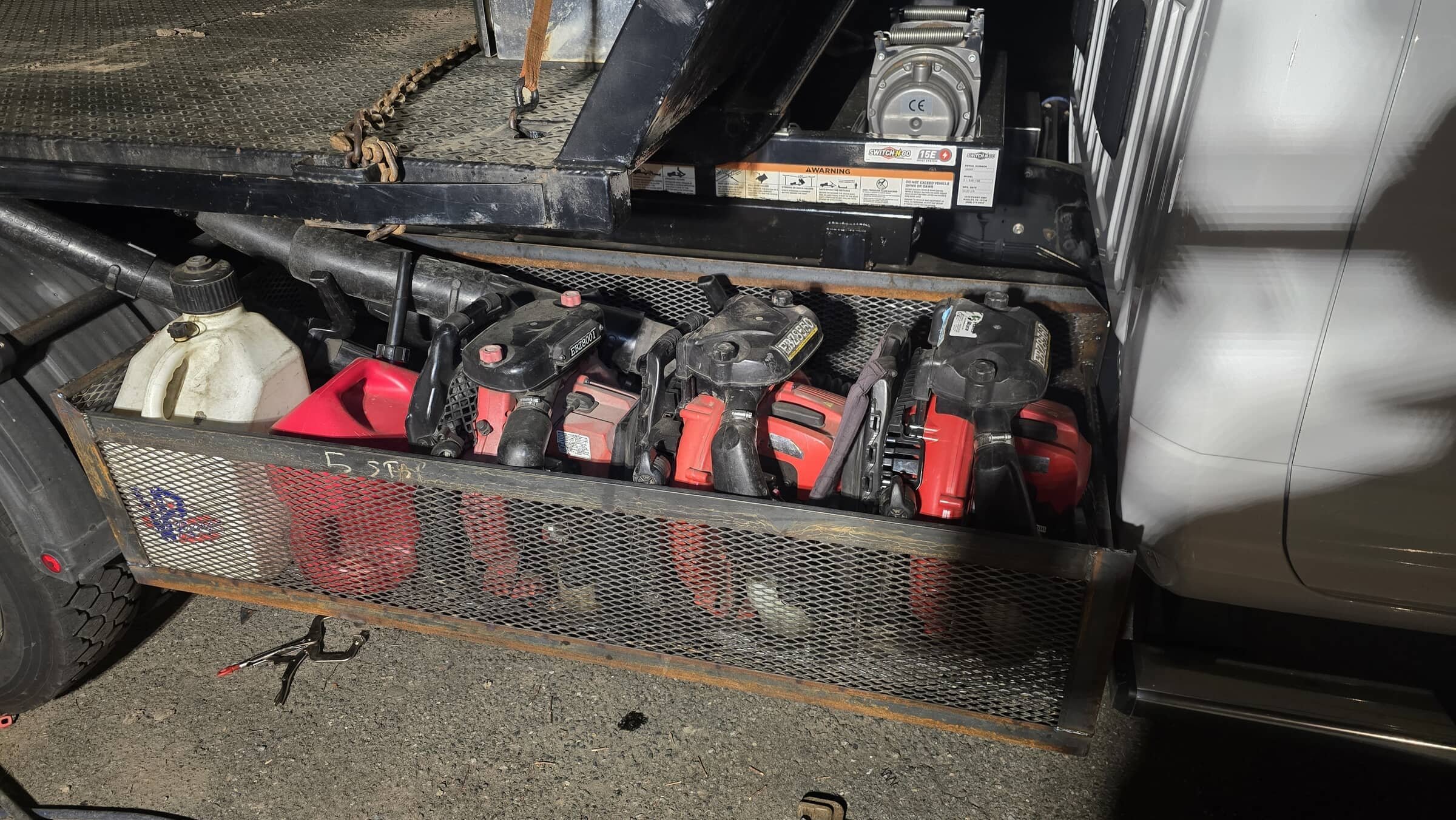 A metal storage container containing power tools, including multiple cordless drills, and a plastic container with a black cap, located on the back of a vehicle.