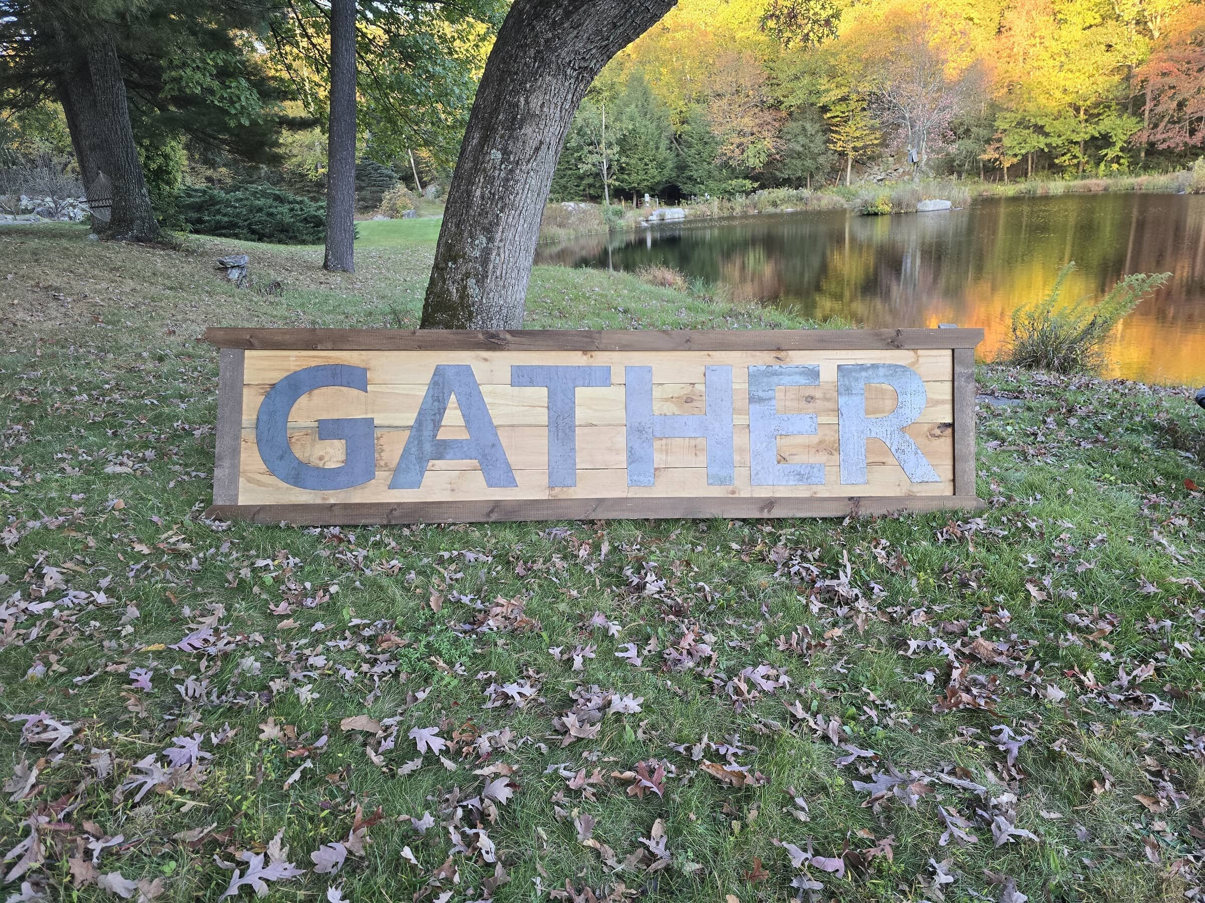 Wooden sign with the word 'GATHER' in large capital letters, placed outdoors on grass near a pond with trees in the background.