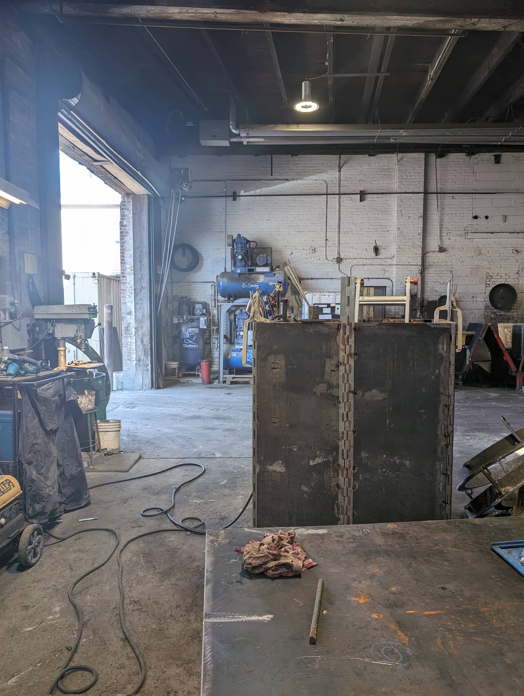 Inside a workshop with various tools and equipment, a large metal box, an open garage door letting in natural light, and a worktable with a rag and a metal rod.
