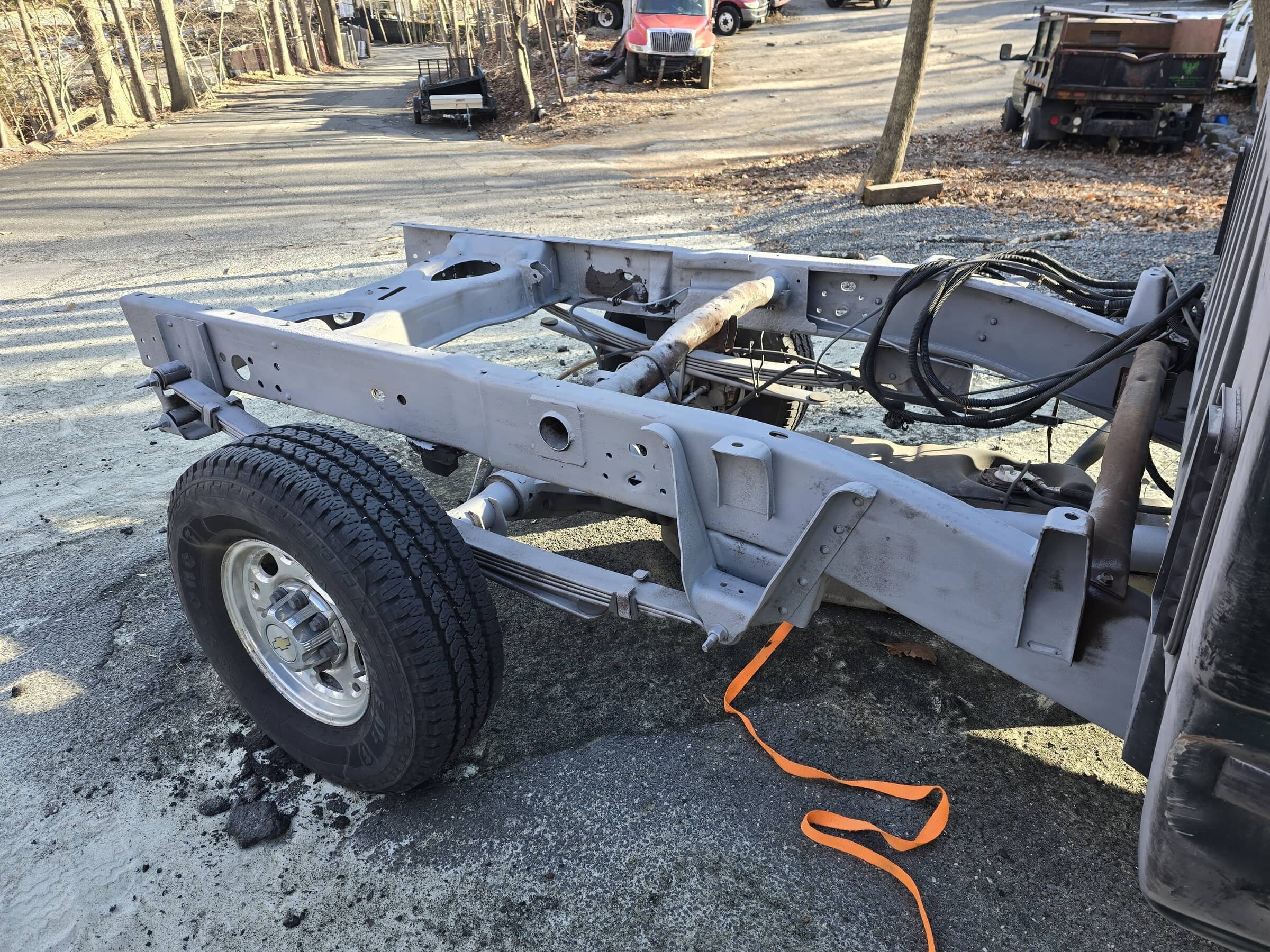Front chassis of a vehicle with one tire and some wires, no engine or body installed, parked on a gravel lot with trees and trucks in the background.