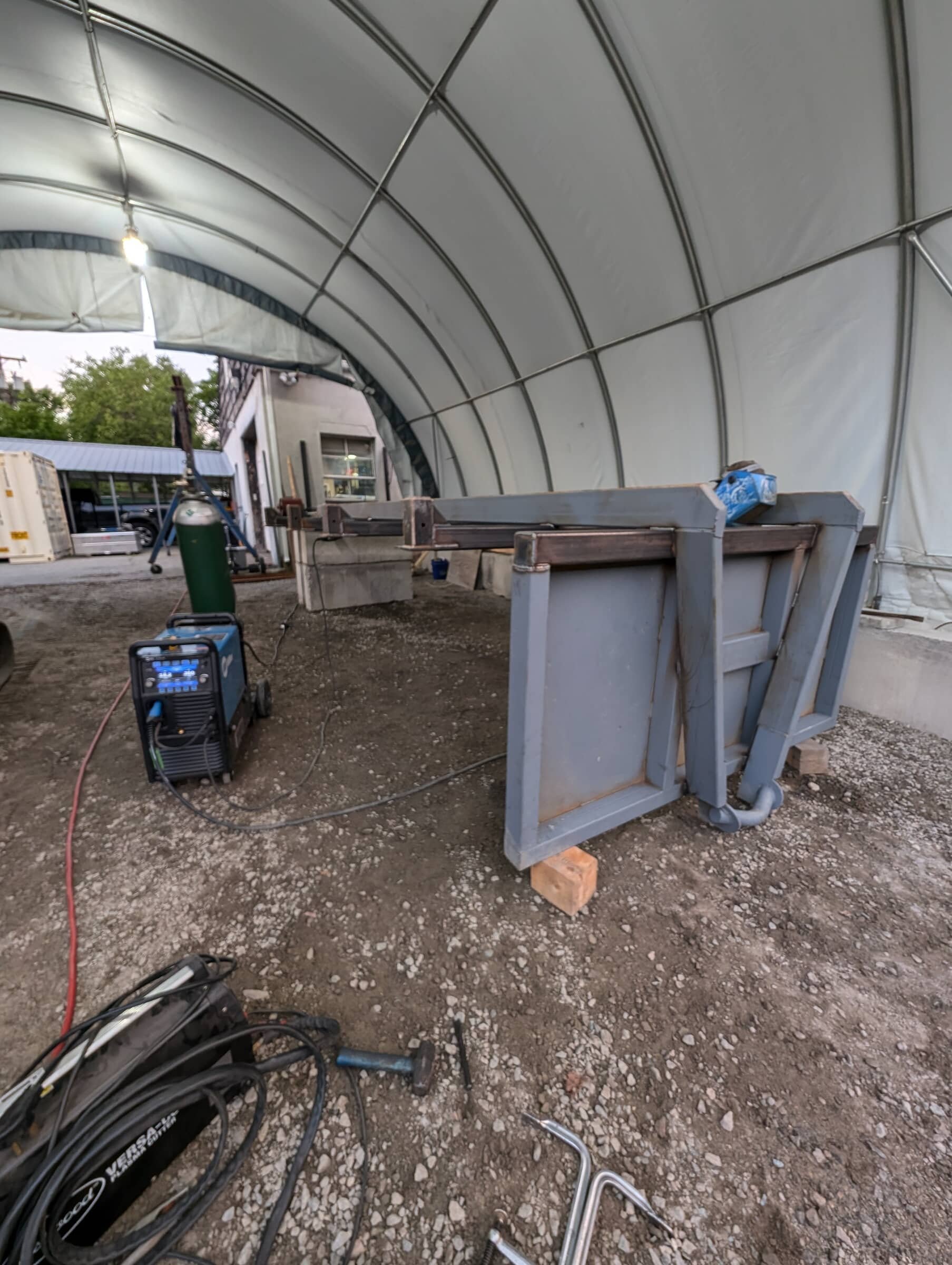Construction site inside a large white tent with metal structures, equipment, and tools on a dirt ground.