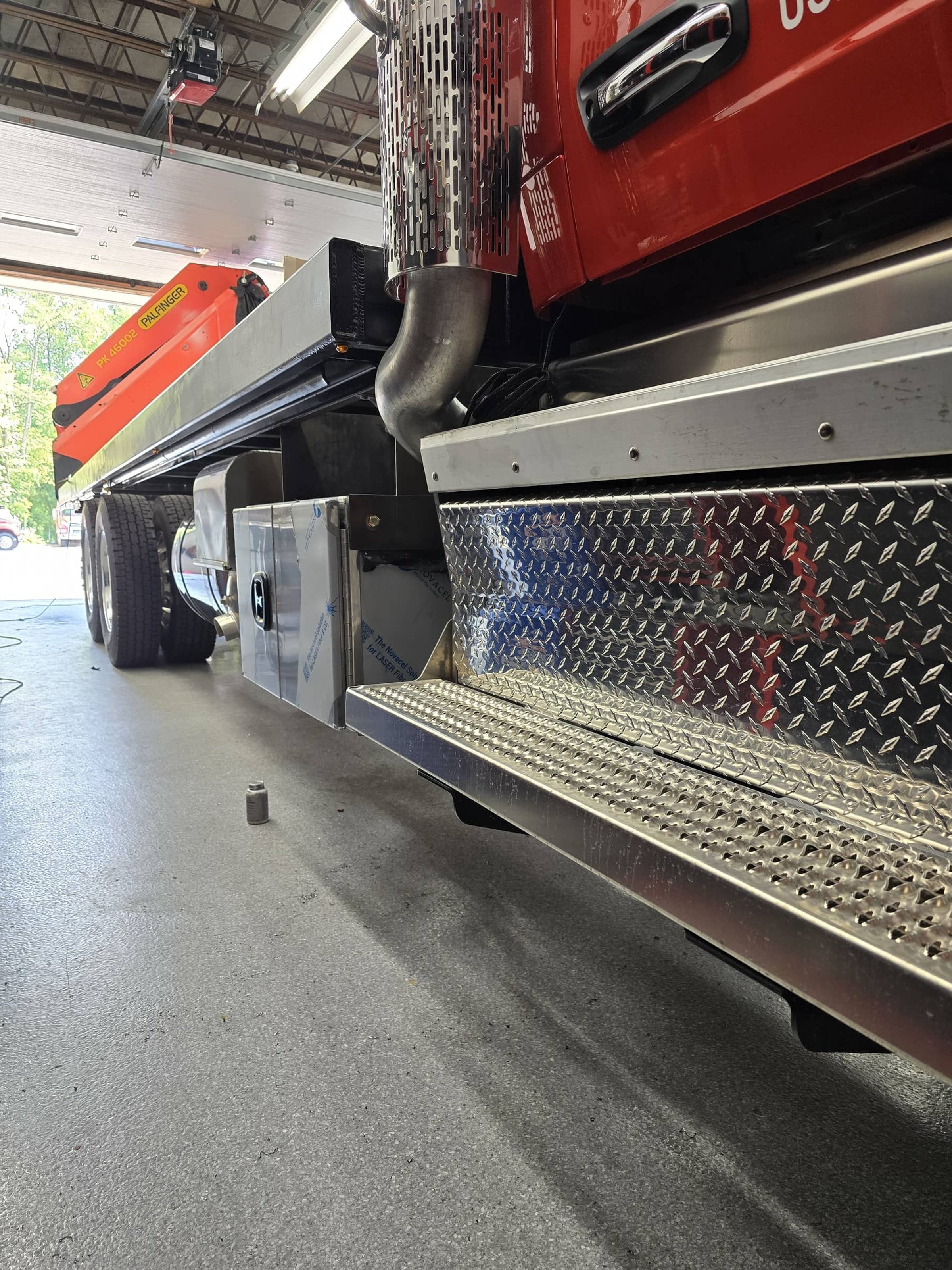 Close-up of the side of a fire truck showing a metal step, storage compartments, and part of the truck's red body with a black handle. A large exhaust pipe is visible near the front of the truck.