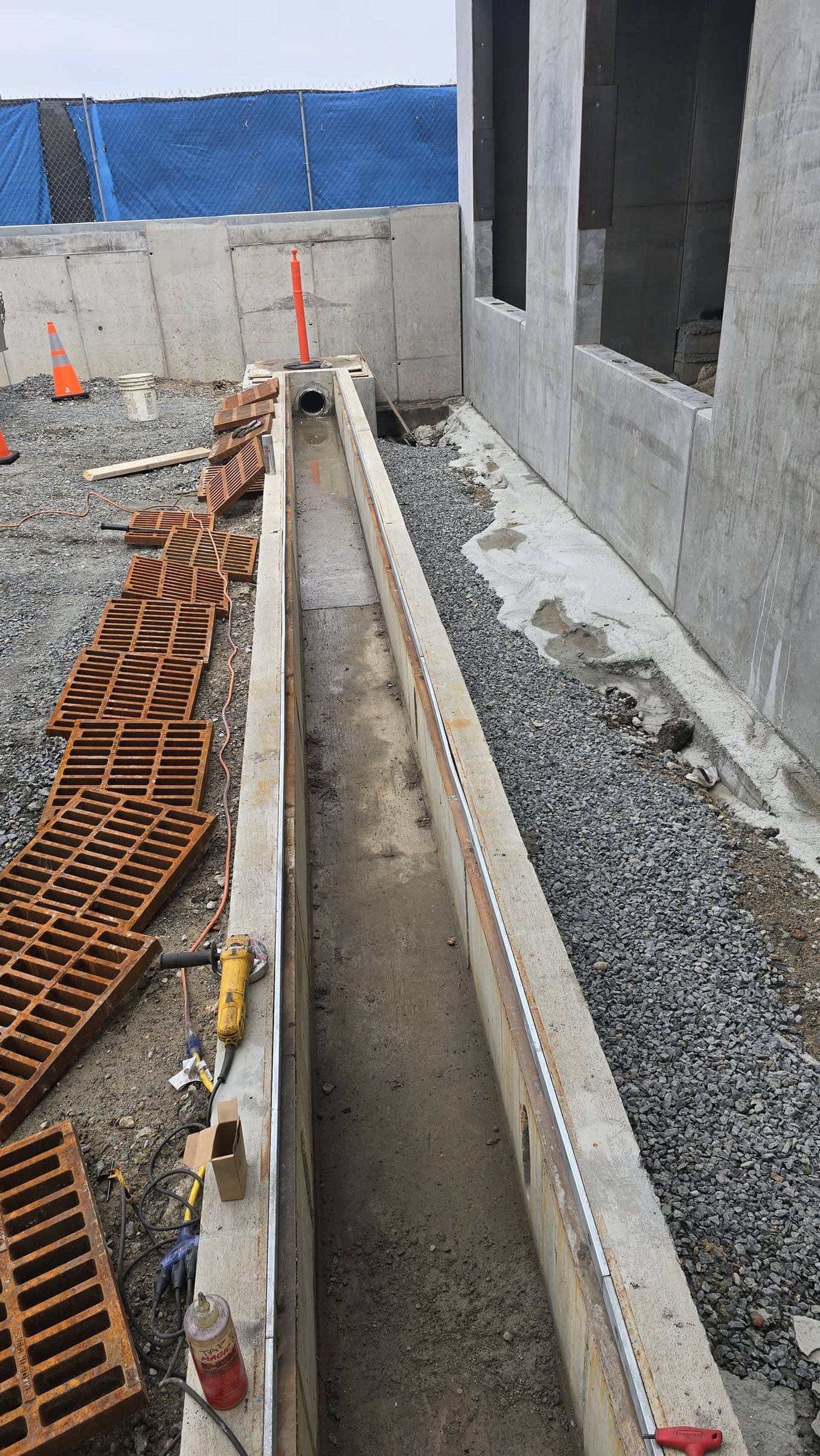 Construction site showing a long, narrow concrete drainage channel with a concrete pipe at the end, surrounded by bricks, construction tools, and gravel.