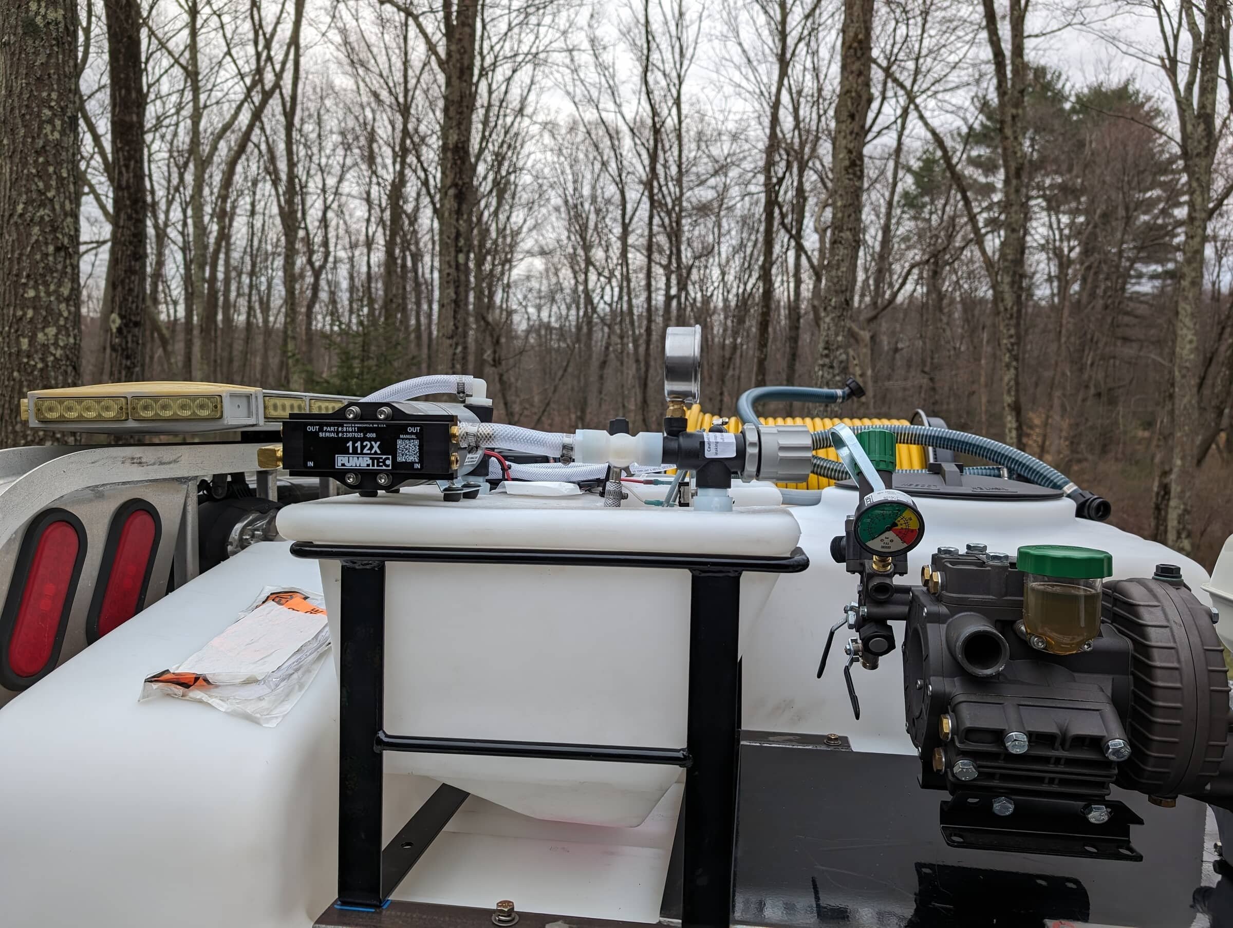 Close-up of a white container with various mechanical components, wires, and gauges on top, set outdoors in a wooded area with leafless trees in the background.