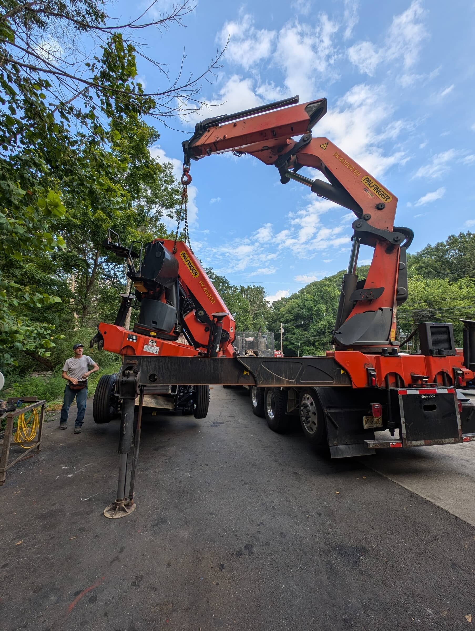 A large orange Palfinger crane truck parked outdoors with a man standing nearby, trees and a partly cloudy sky in the background.