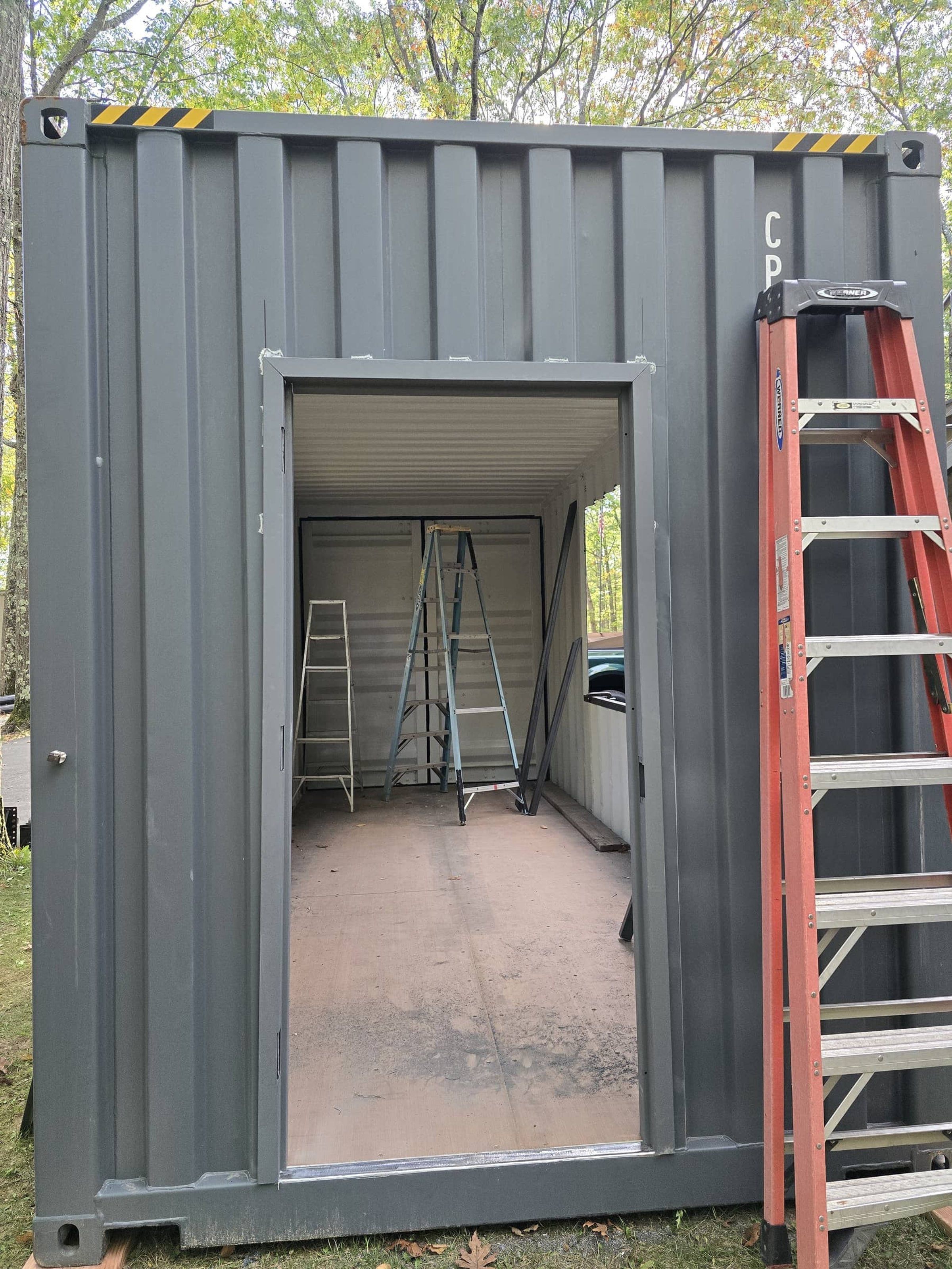 The image shows a partially constructed small building made from an upcycled shipping container, with an open doorway, several ladders inside, and a red step ladder leaning against the right side of the container.