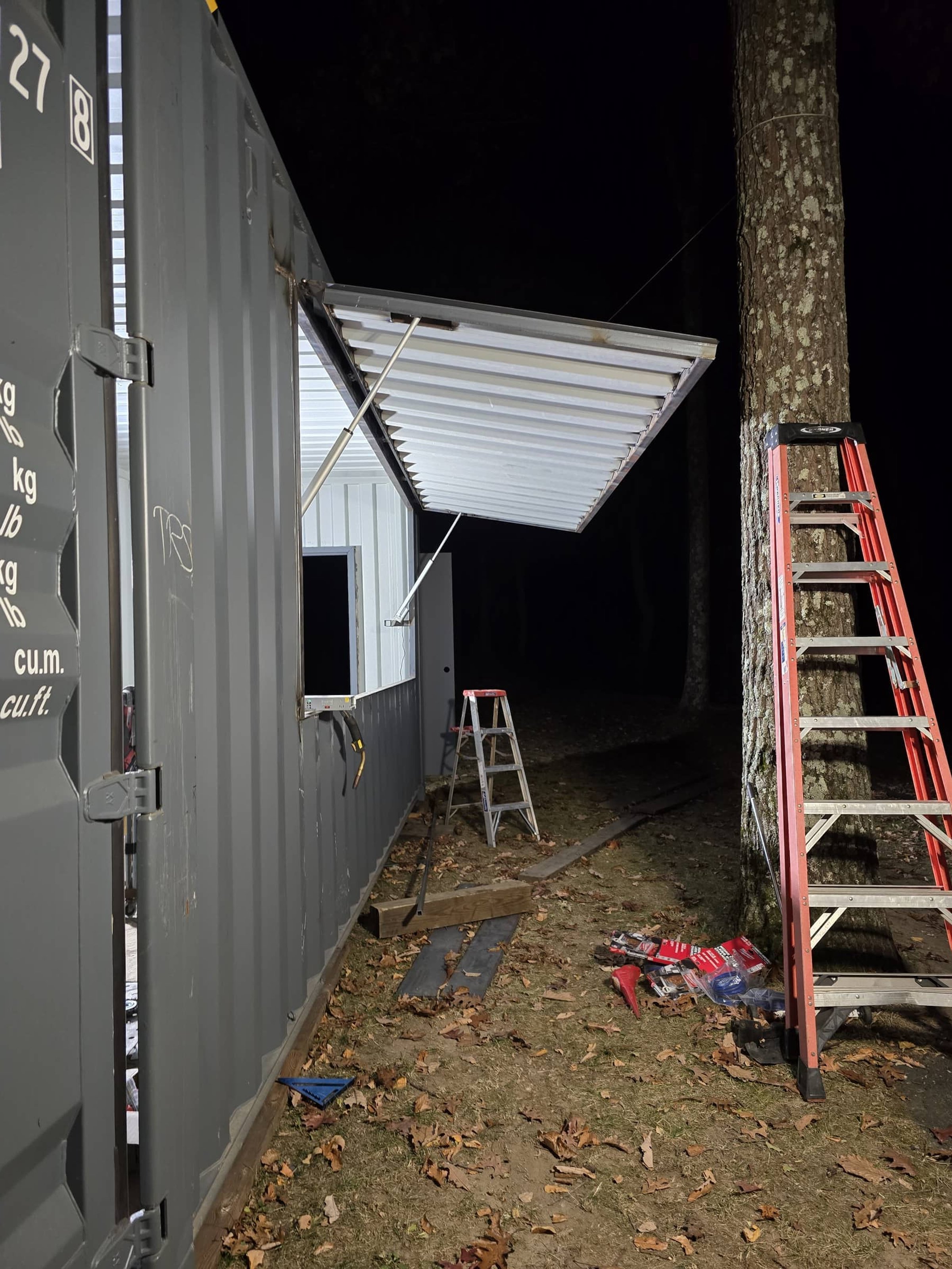 Nighttime outdoor scene with a partially installed metal awning extending from a gray building, a red ladder leaning against a tree, and various tools and debris on the ground.