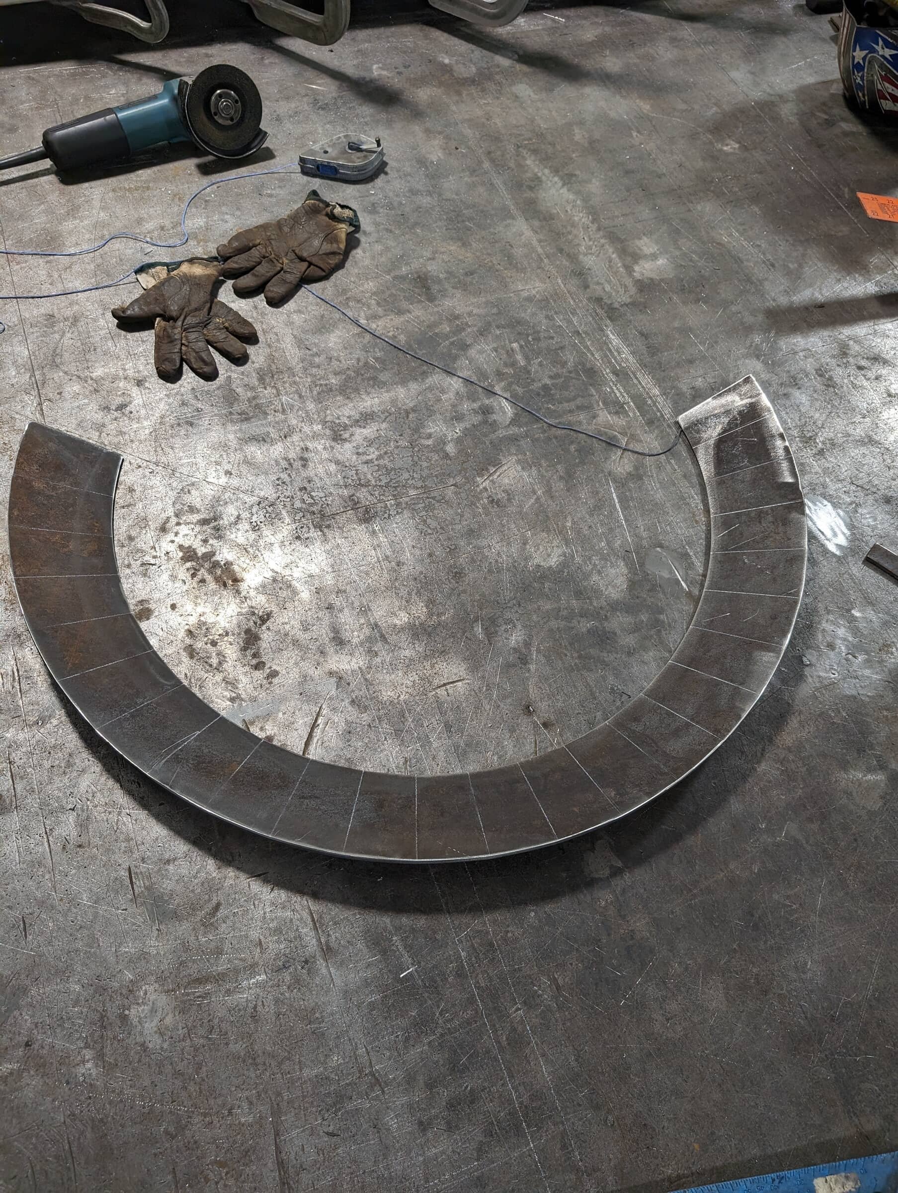 A large curved metal piece on a workshop floor, surrounded by safety gloves, a grinder, and various tools.