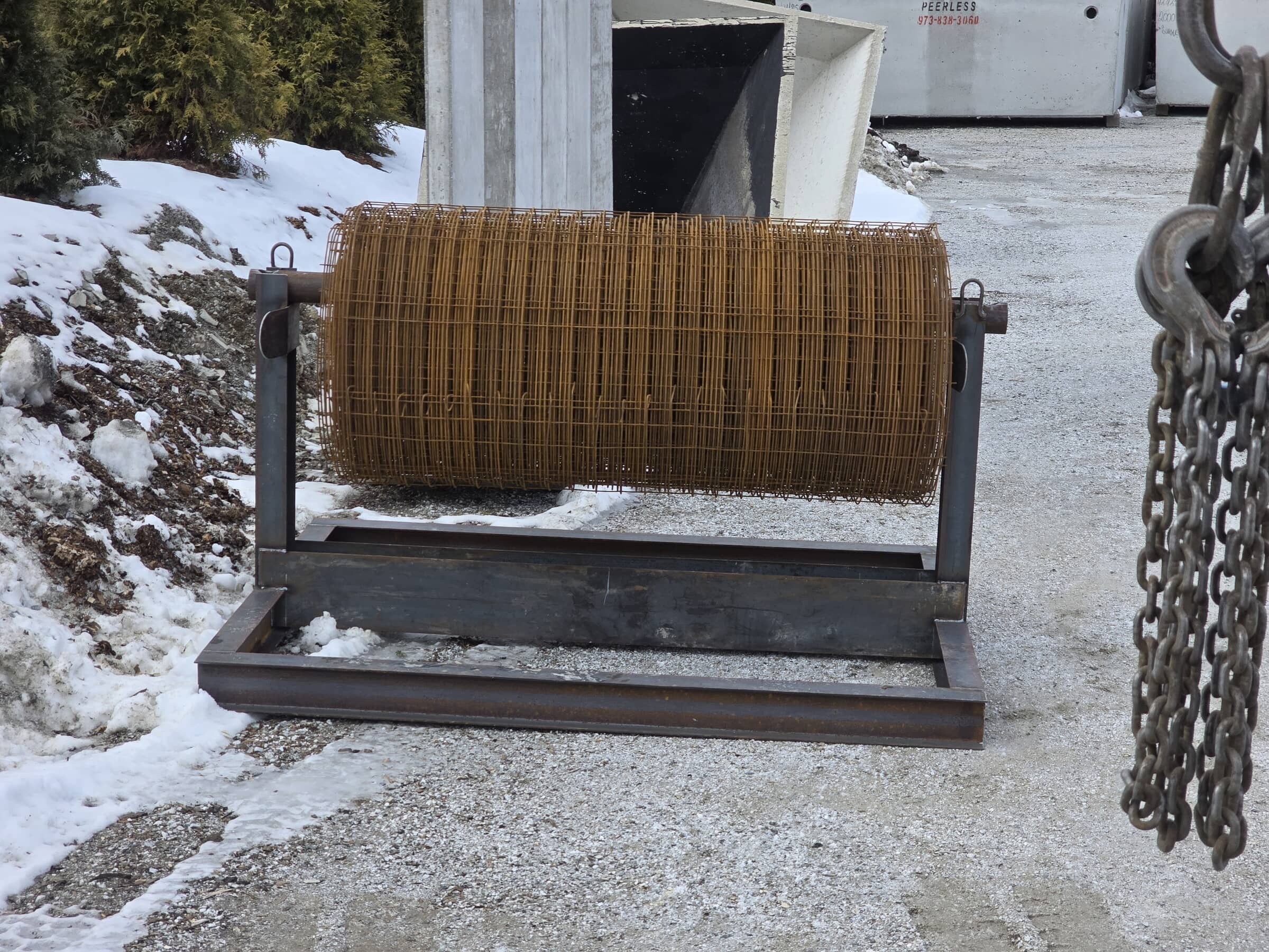 A metal frame with a large cylindrical wire mesh roll on it, outdoors with snow on the ground, surrounded by chains and industrial equipment.