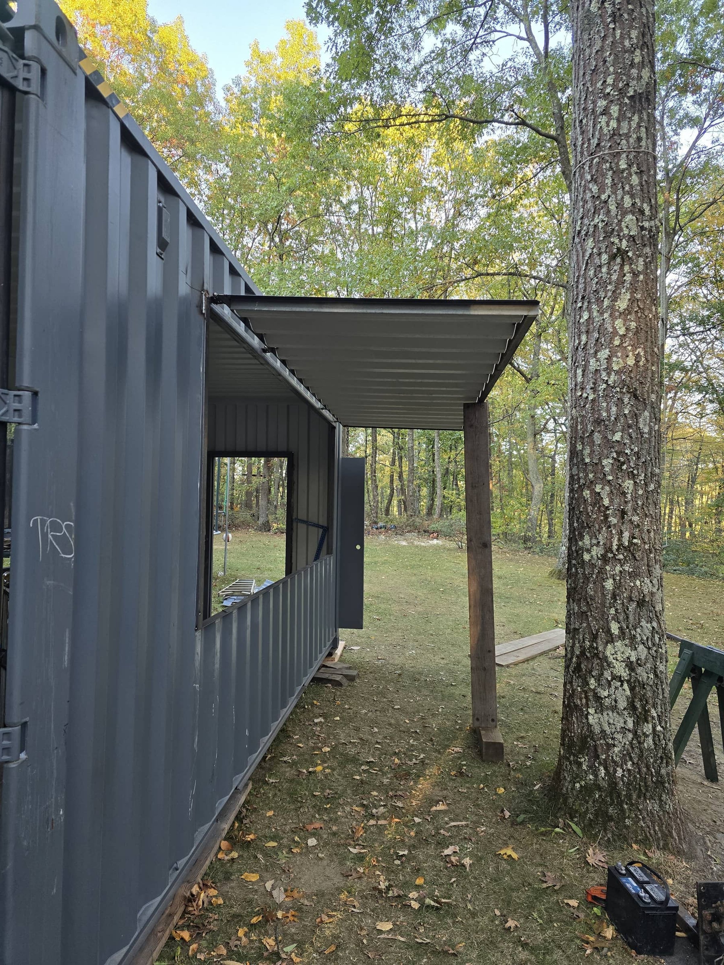 View of a metal container with a window and open door, situated outdoors among trees, with an extension made of wood supports and metal roofing. There are scattered leaves on the ground.