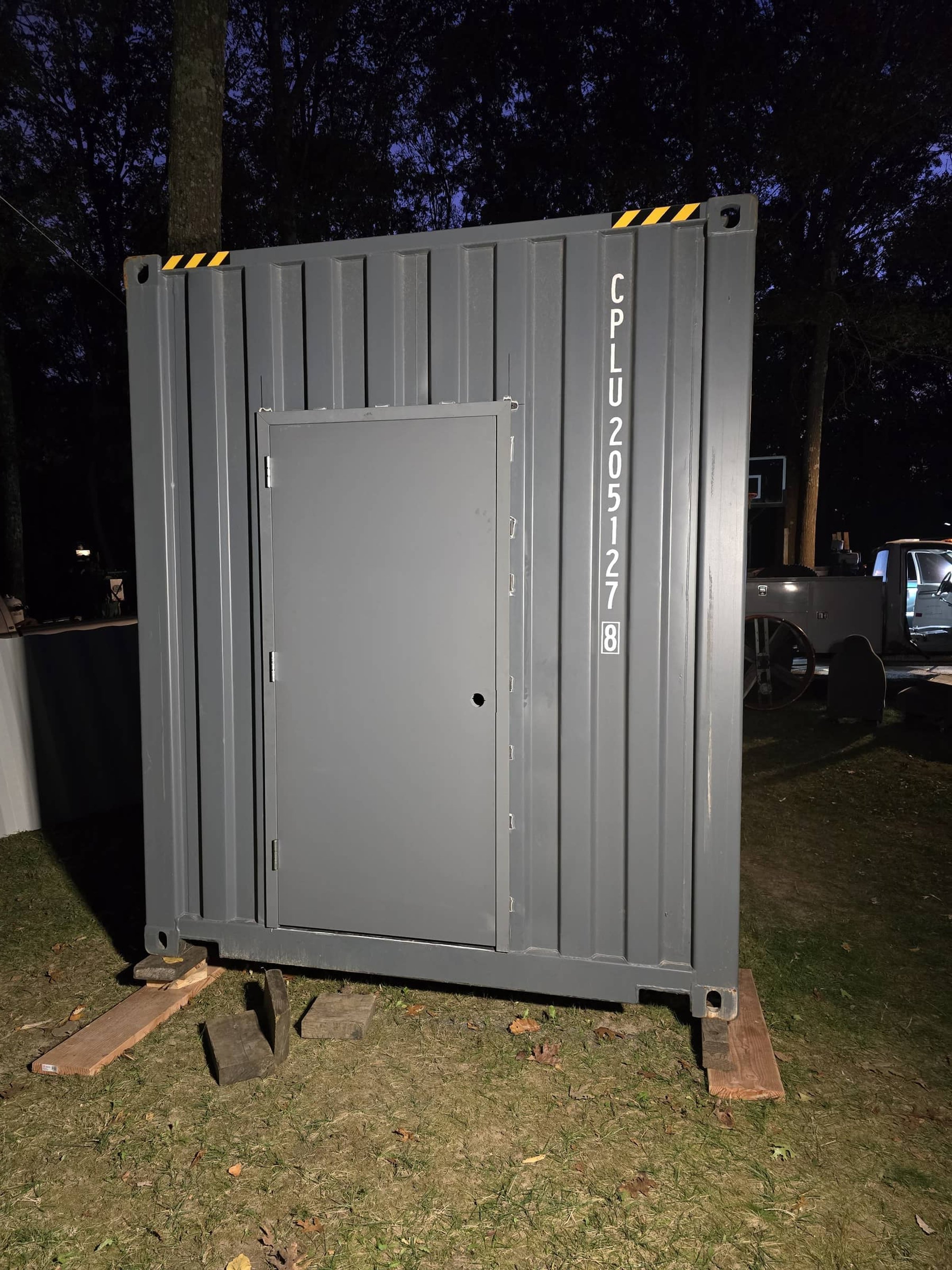 Gray metal shipping container with a door, placed on wooden blocks and outdoors at dusk, surrounded by trees and other equipment.