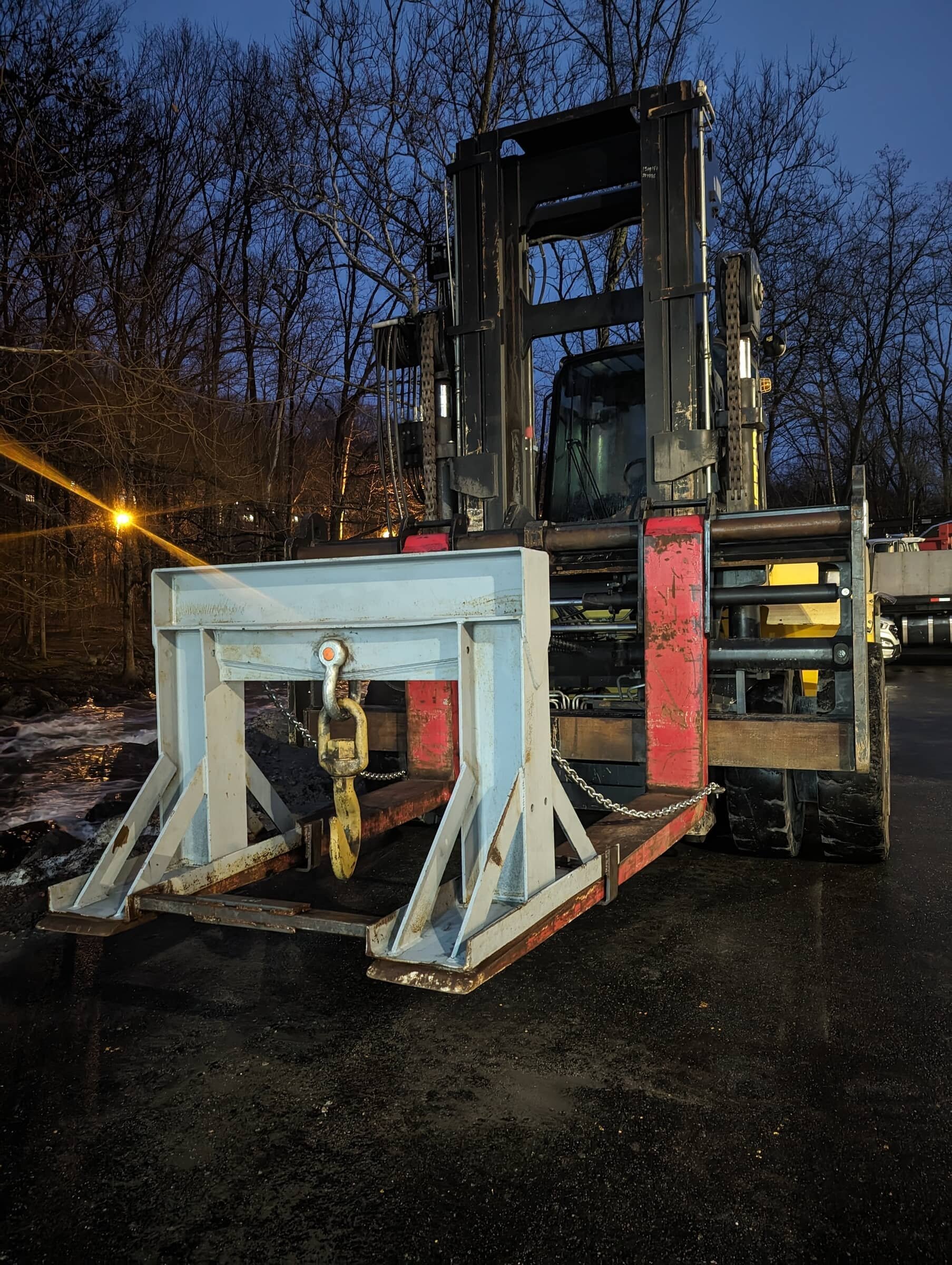 A forklift truck is parked on a dark, wet surface with a steel attachment on its front, set against a background of leafless trees during dusk or dawn.