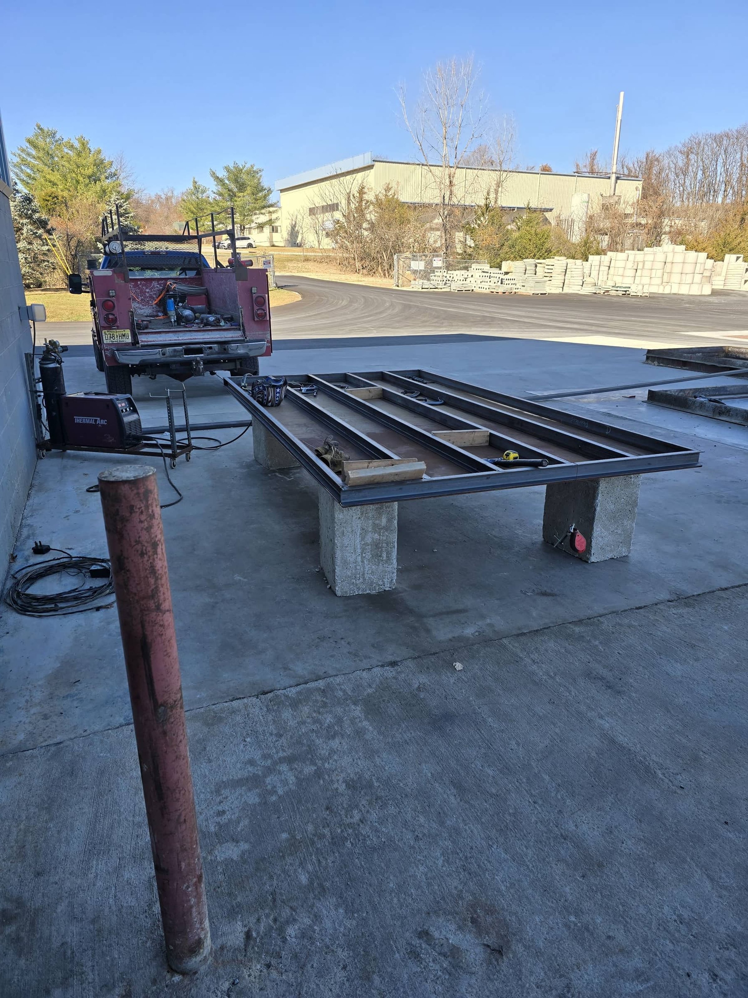 Construction site with metal frame on cinder blocks, red truck, tools, and industrial supplies, under clear blue sky.