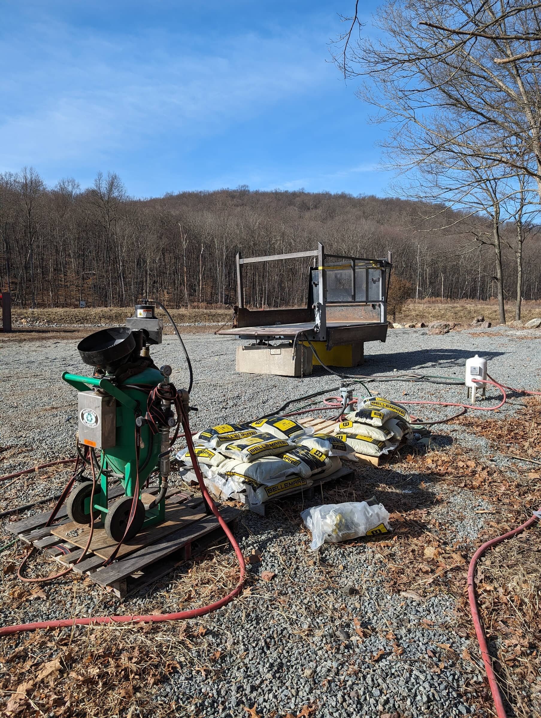 Construction site with equipment, bags of material, and a small platform on gravel outdoors, with leafless trees and a hill in the background.