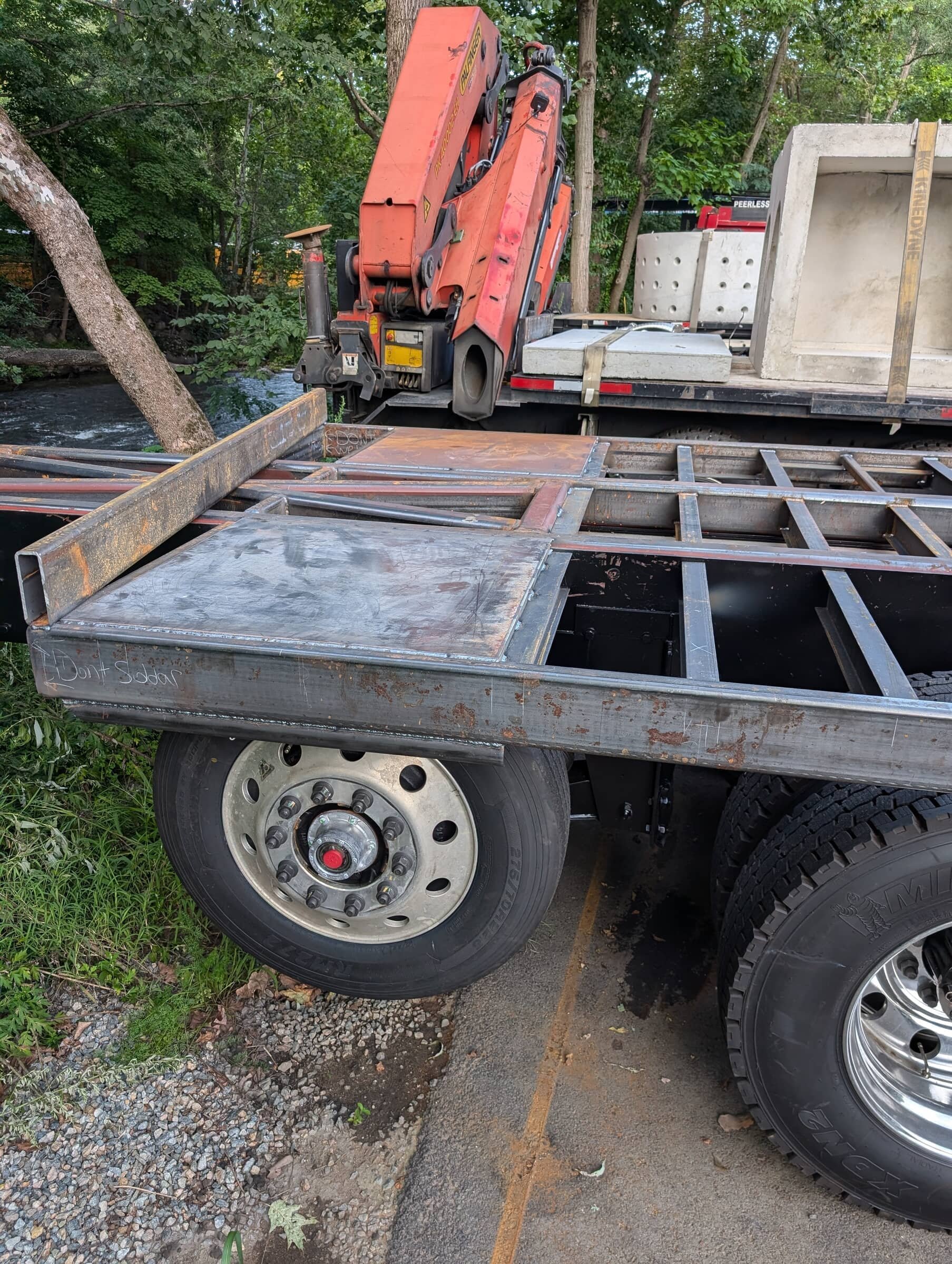 Empty metal flatbed truck with a mounted crane, parked near a wooded area and a river.