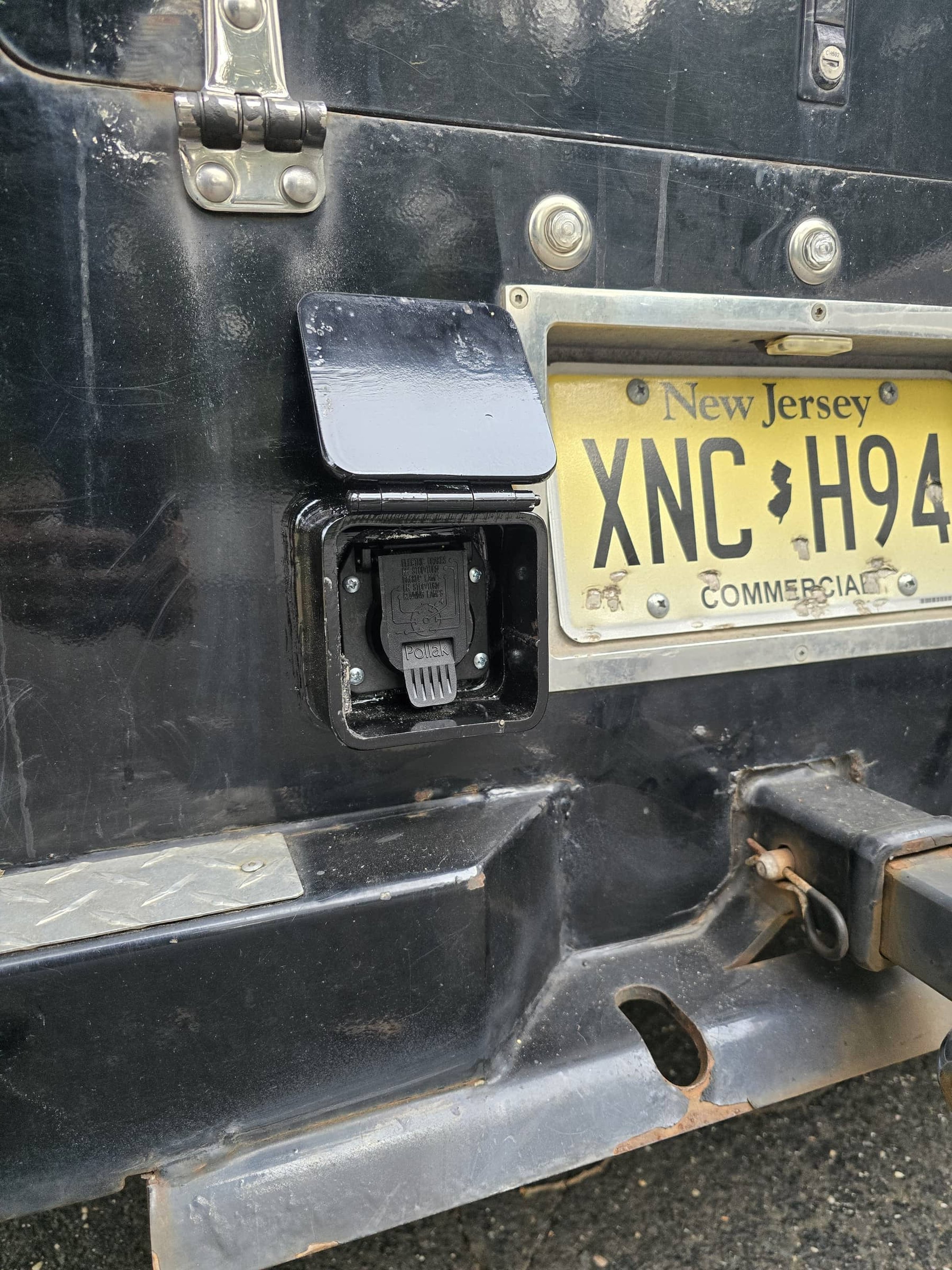 Close-up of a New Jersey license plate on a vehicle with a black hitch receiver and electrical connection port below the license plate.