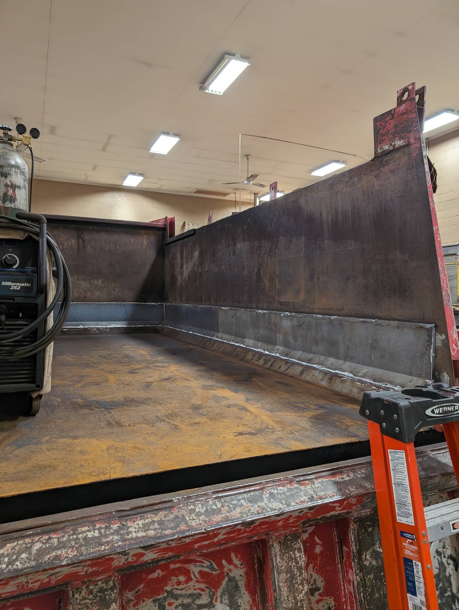 The image shows the bed of a work truck with a welded steel side panel, a Millermatic 252 welding machine on the left, and an orange Werner step ladder on the right, in a workshop with fluorescent lighting.