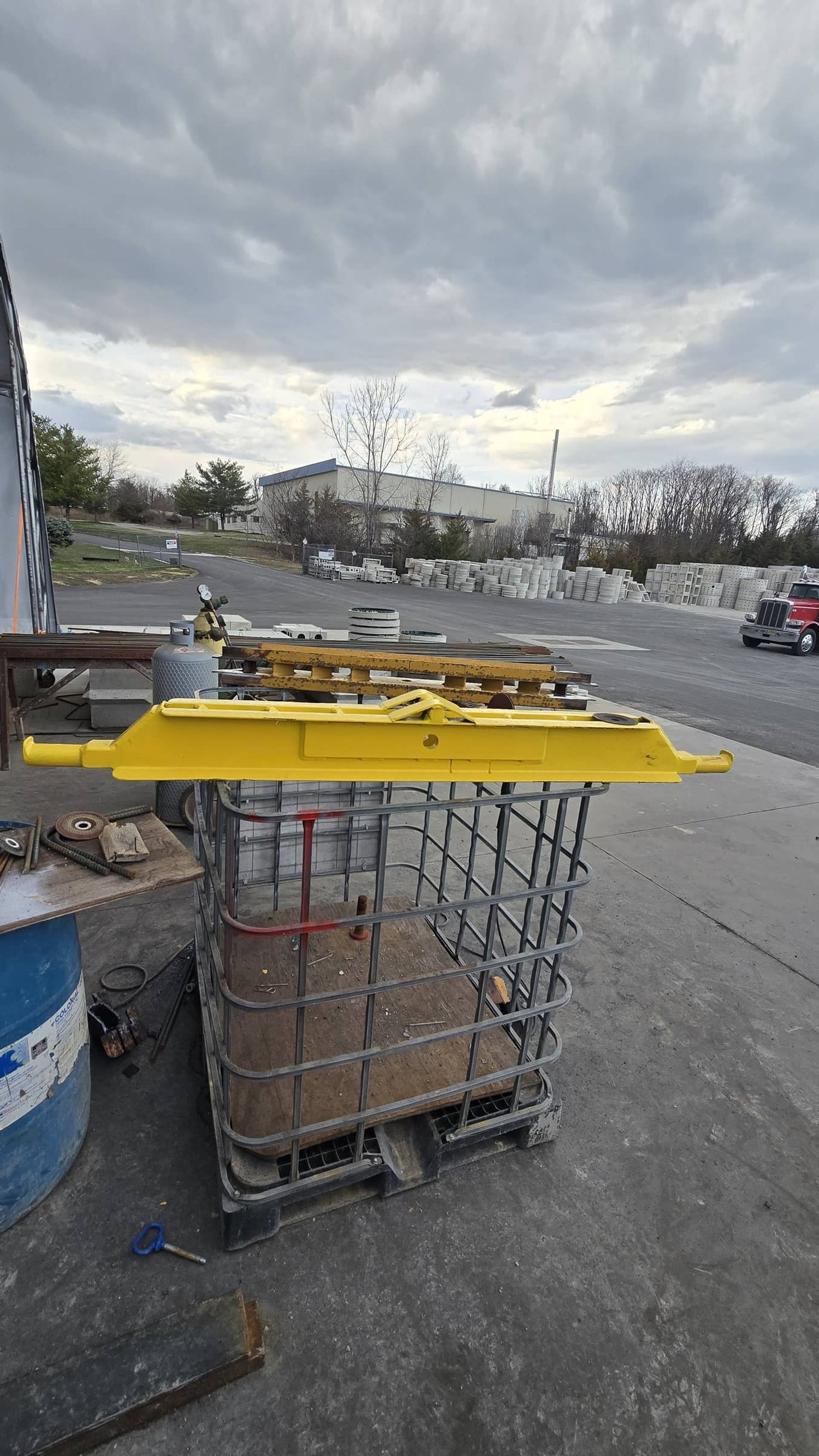 Outdoor industrial workspace with a yellow plastic tool resting on a wire storage bin, surrounded by miscellaneous tools and equipment, with a cloudy sky overhead.